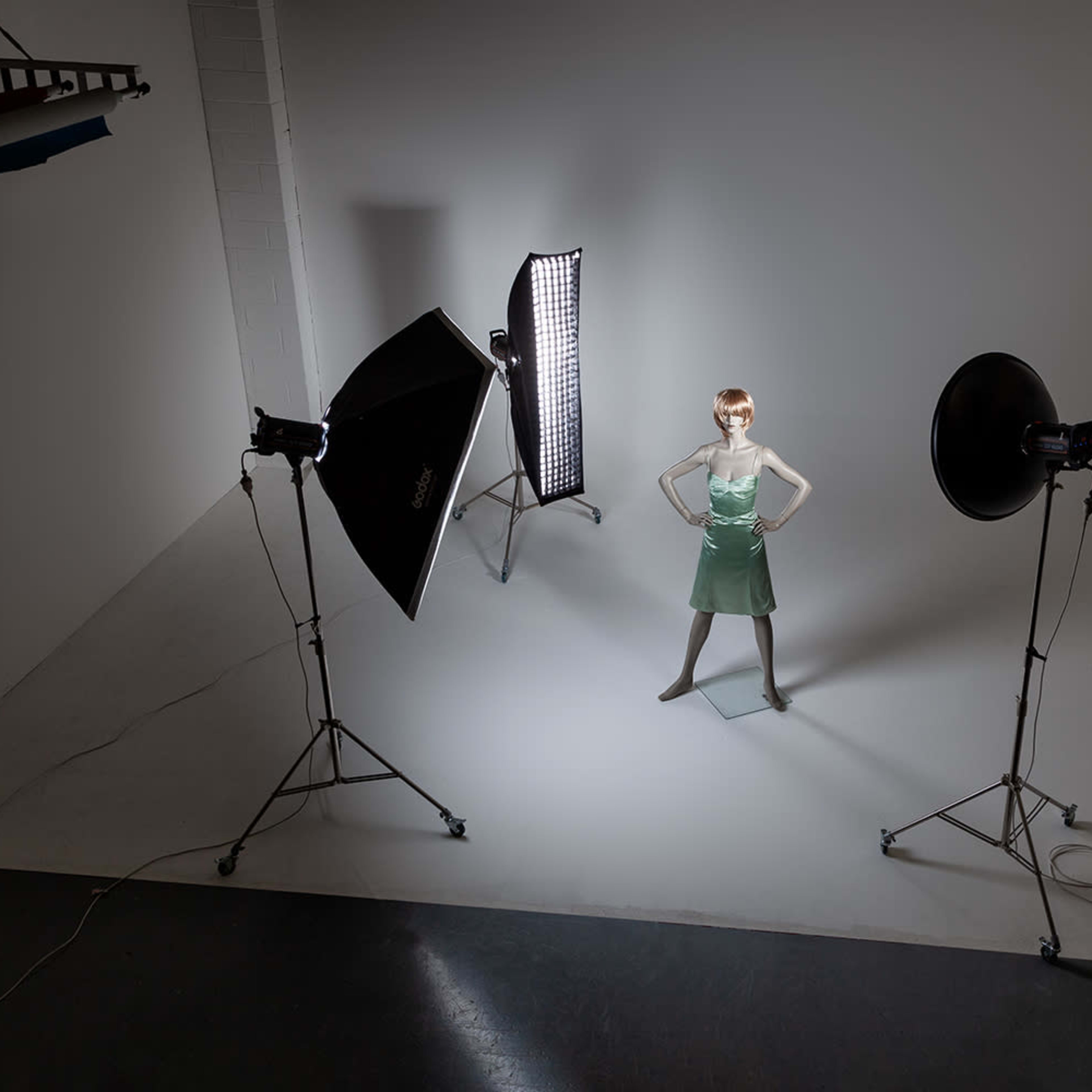 A mannequin dressed in a green dress stands in the center of a photography studio surrounded by lighting equipment.