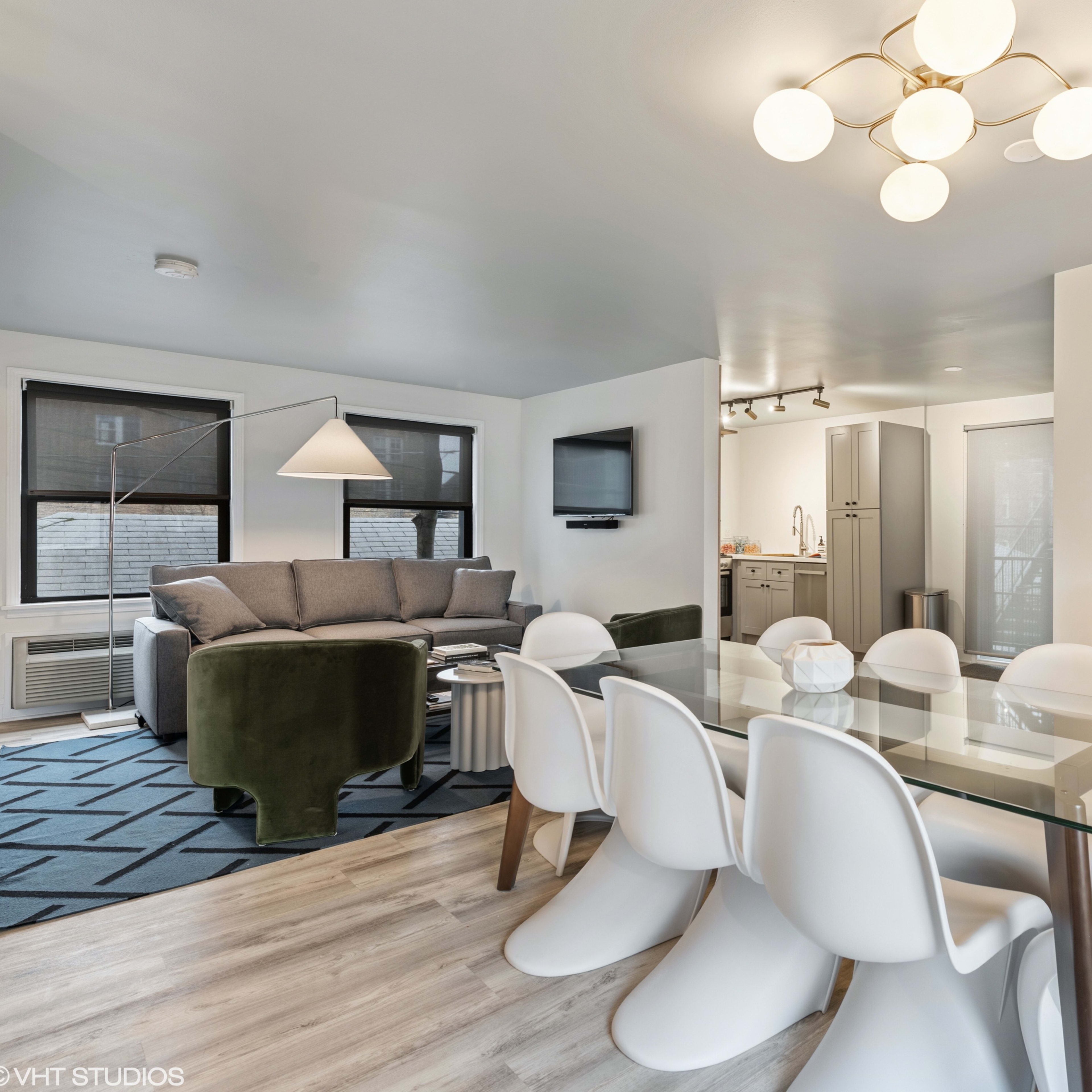 A modern living space featuring a gray sofa, a dining table with white chairs, and a kitchen area in the background.