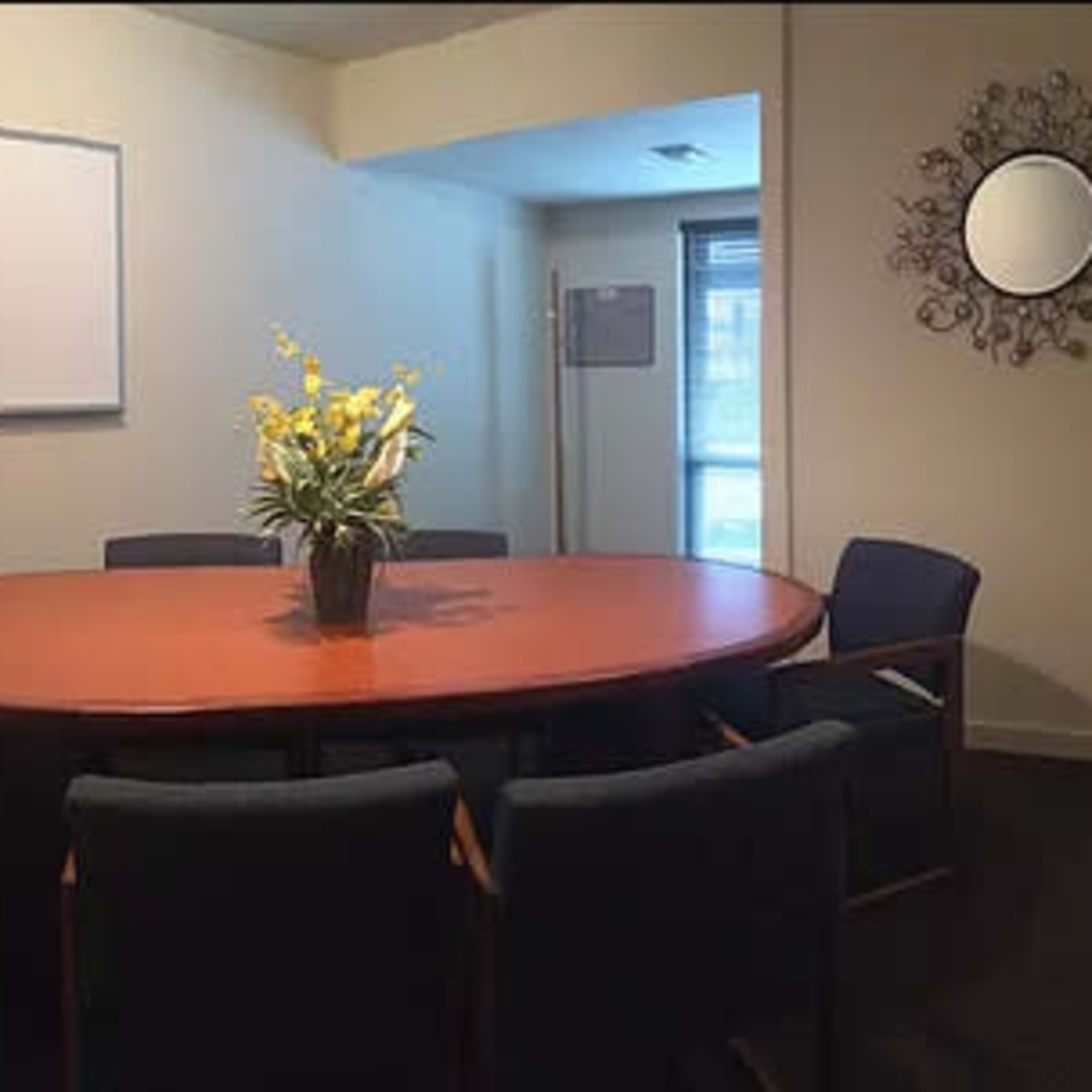 A conference room features a large oval table surrounded by four chairs, with a potted plant in the center and a whiteboard on the wall.