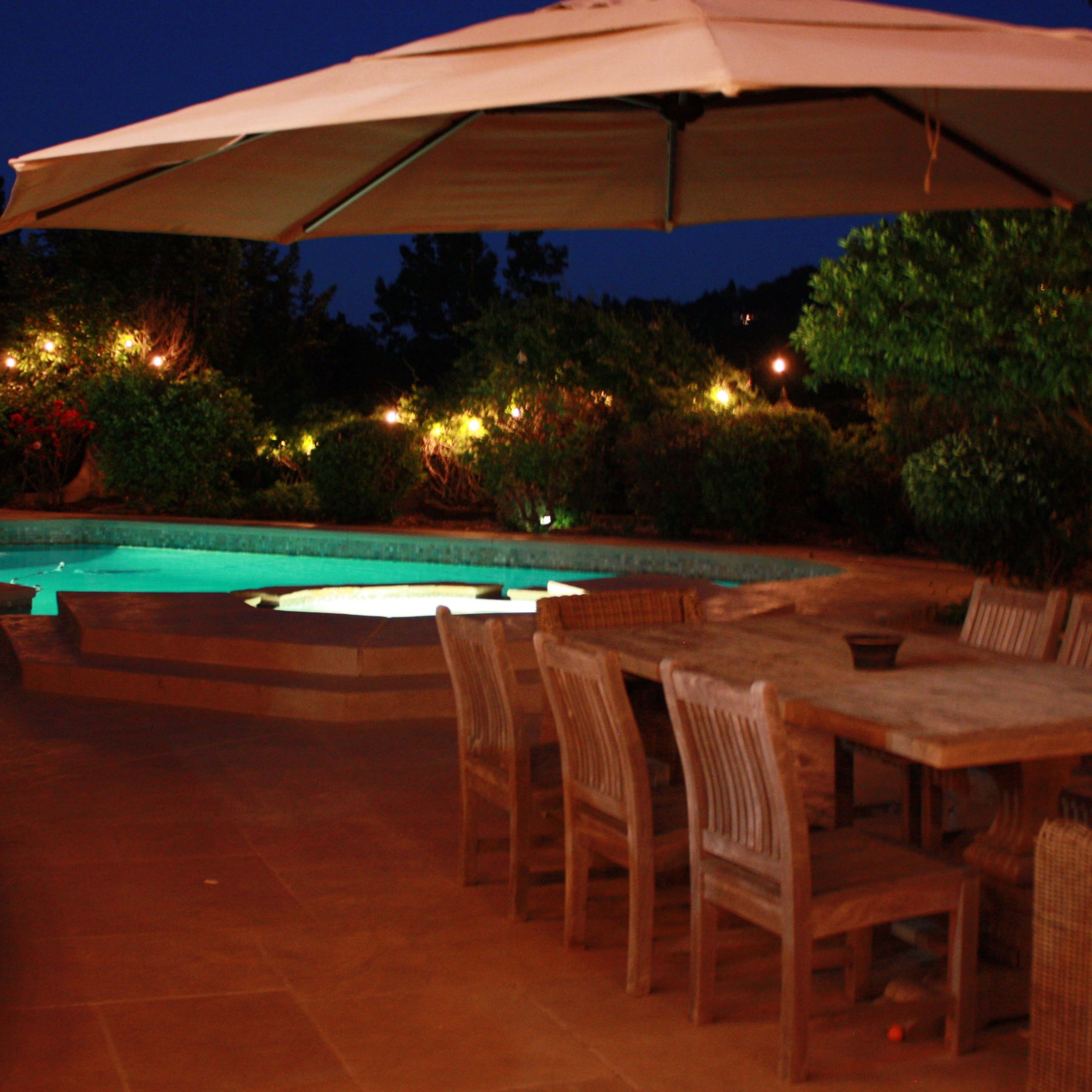 A patio area features a wooden dining set and an umbrella beside a lit pool and landscaped greenery at night.
