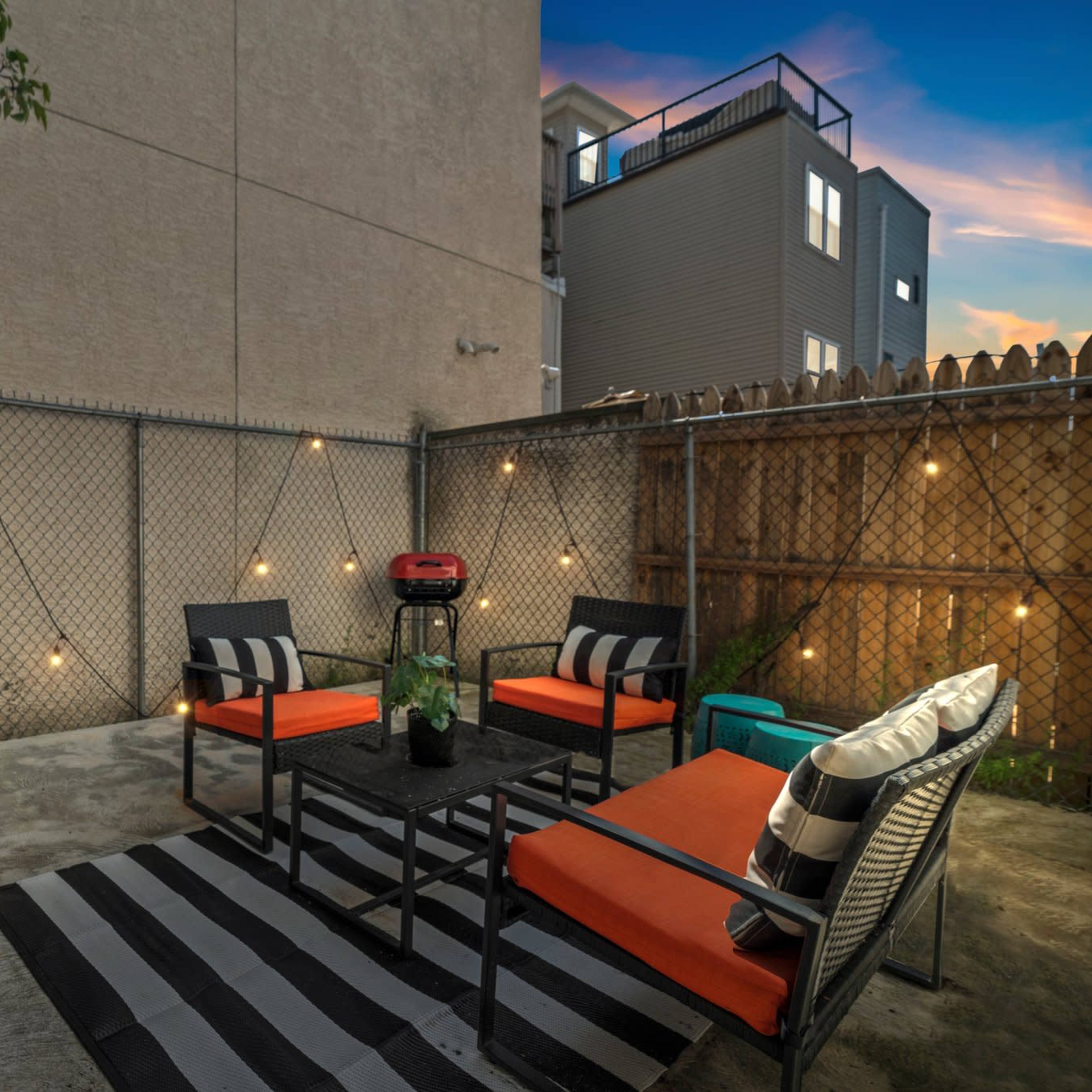 A small outdoor seating area with black and orange furniture, a small grill, and string lights along a chain-link fence.