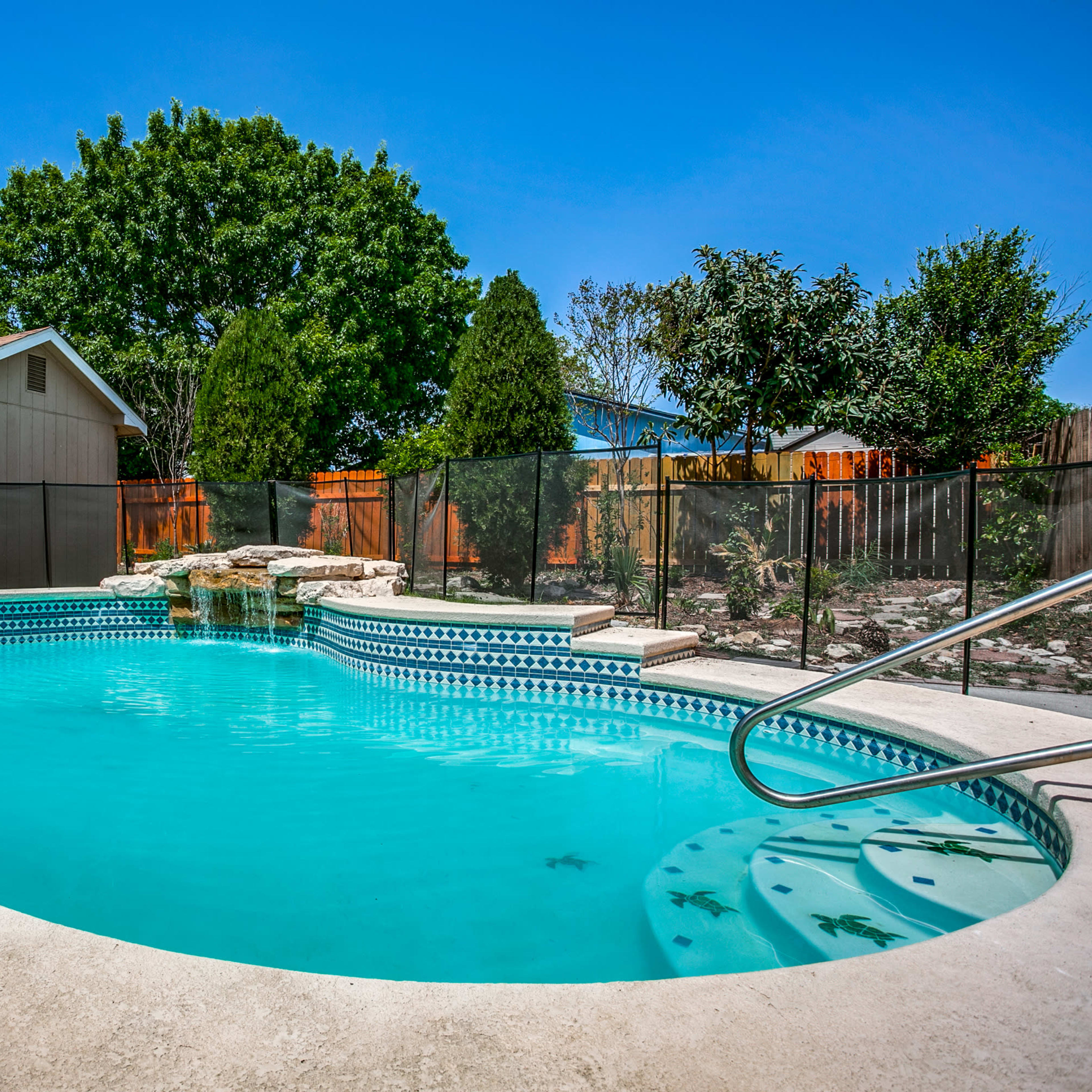 A backyard swimming pool with a waterfall feature, surrounded by a fence and a small shed in the background.