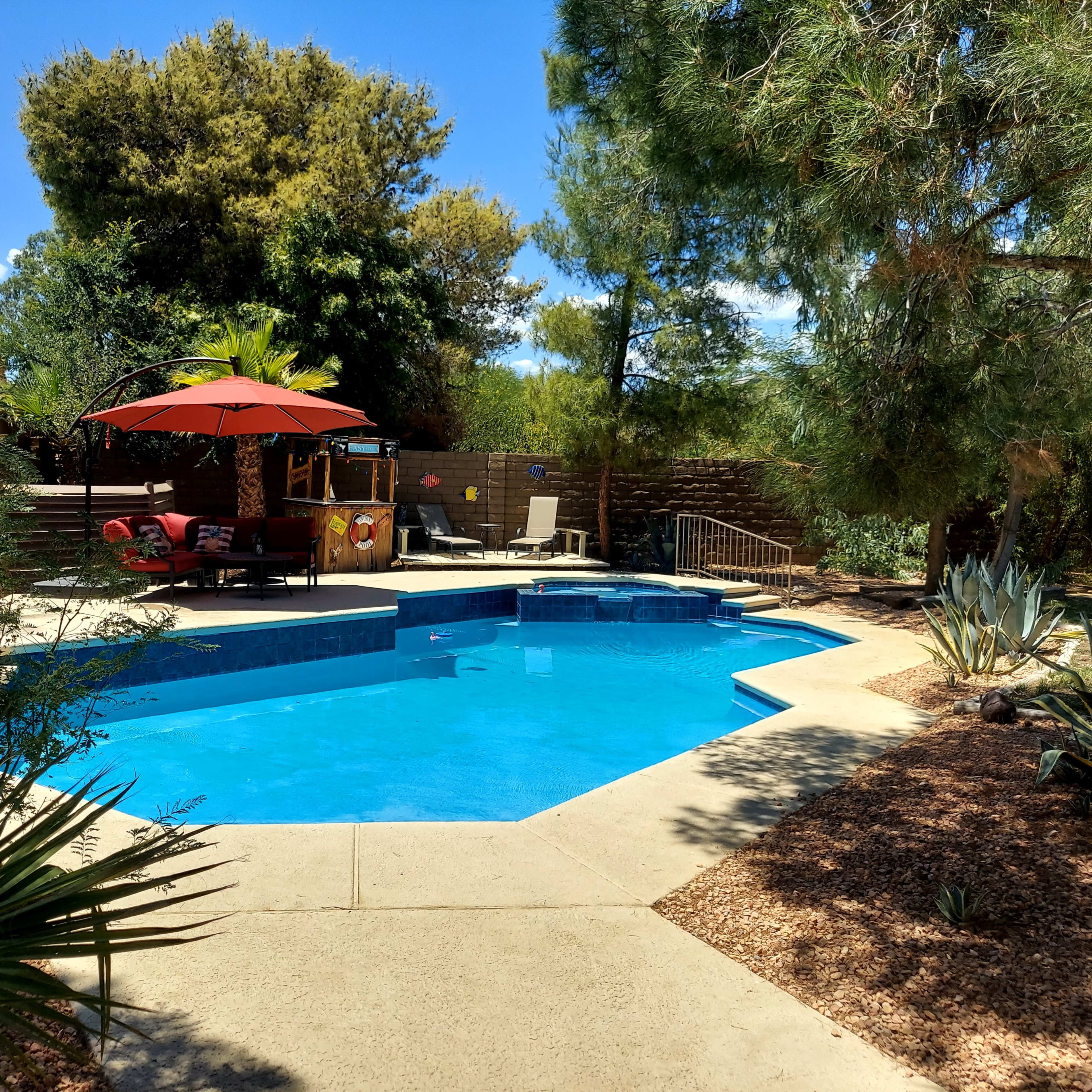 A pool surrounded by a landscaped backyard featuring palm trees, lounge chairs, and an umbrella.