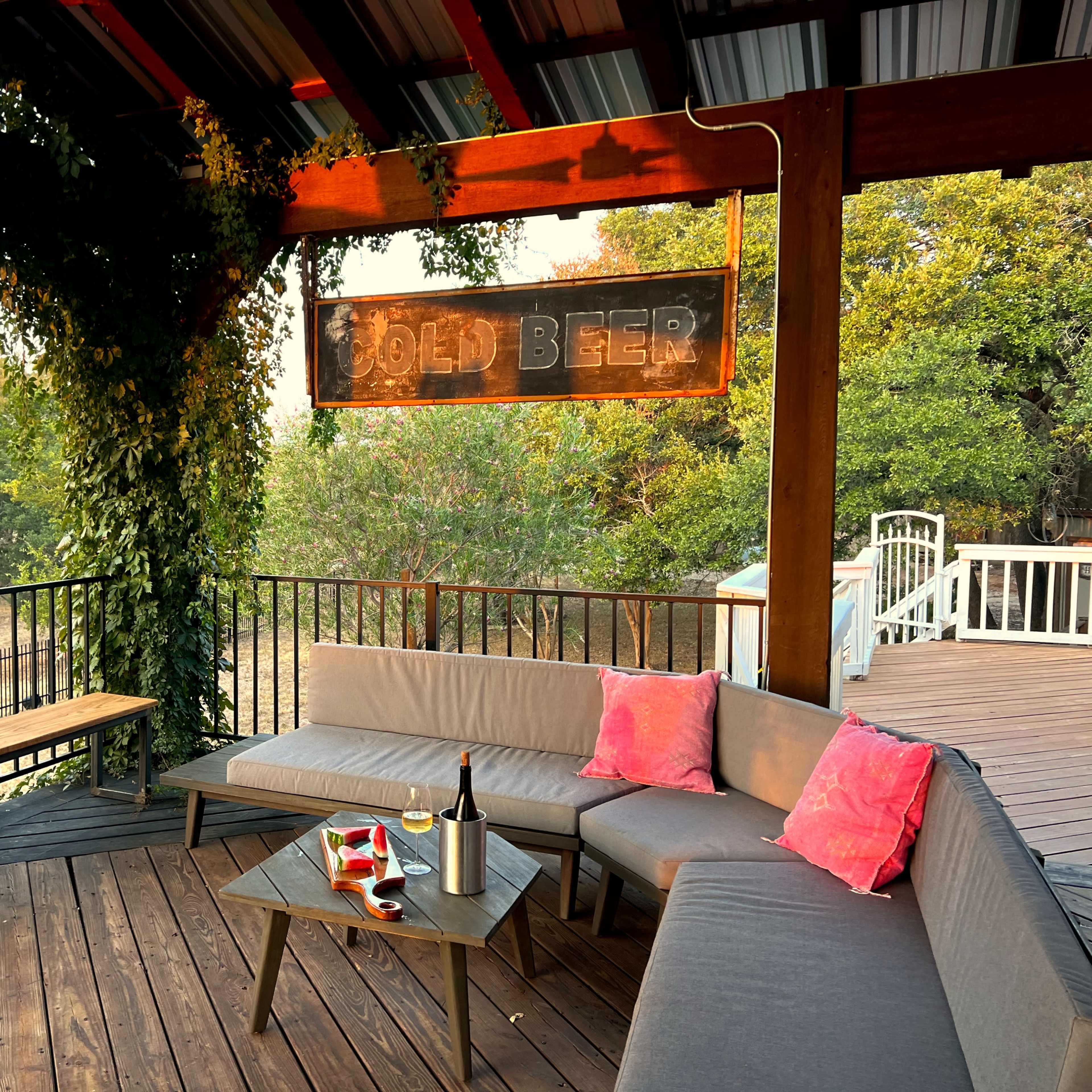A covered outdoor patio features a lounging area with gray sectional seating, bright pink pillows, a small table, and a sign that reads "Cold Beer."