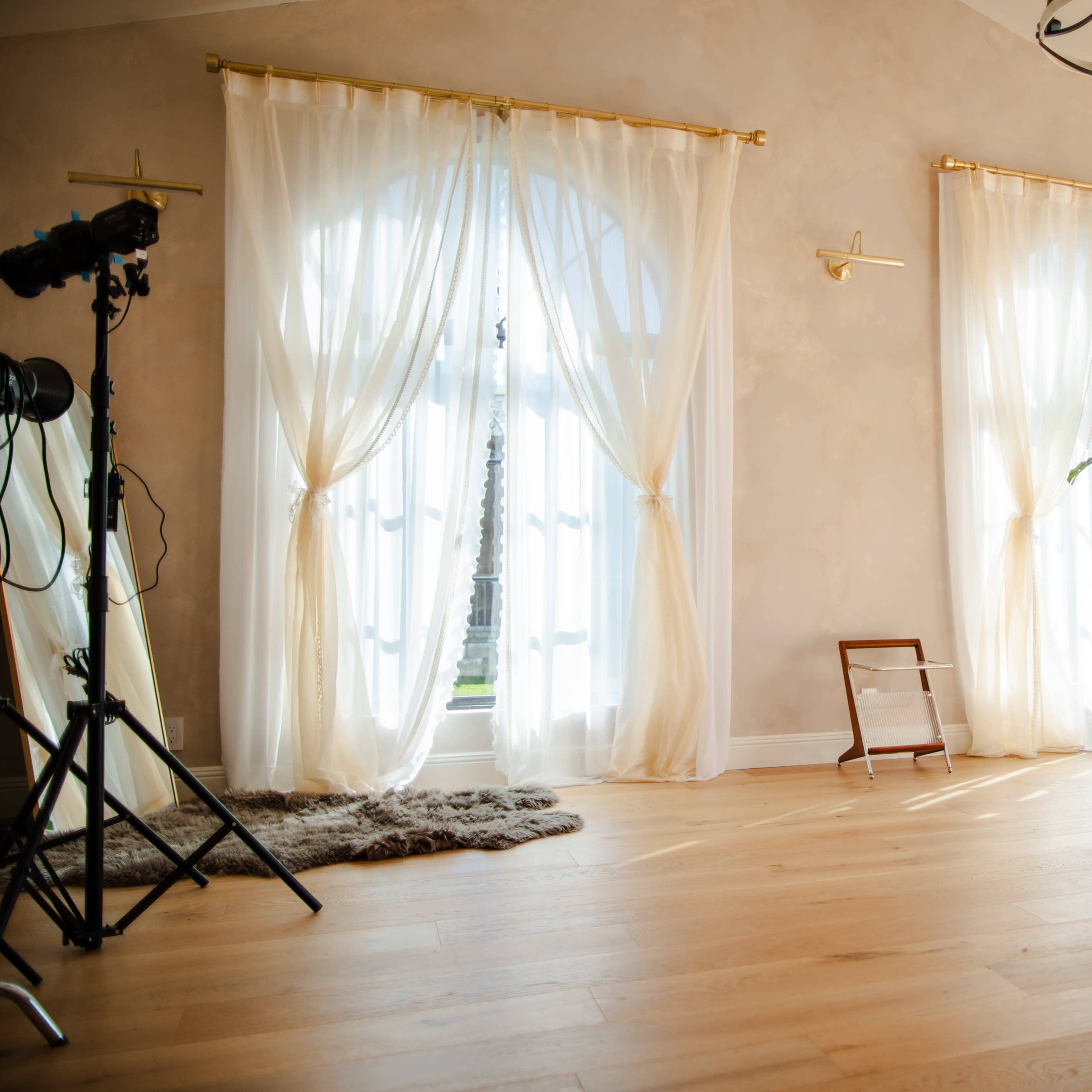 A well-lit room with large windows dressed in sheer curtains, a potted plant, and a minimalist chair, featuring warm wood flooring.