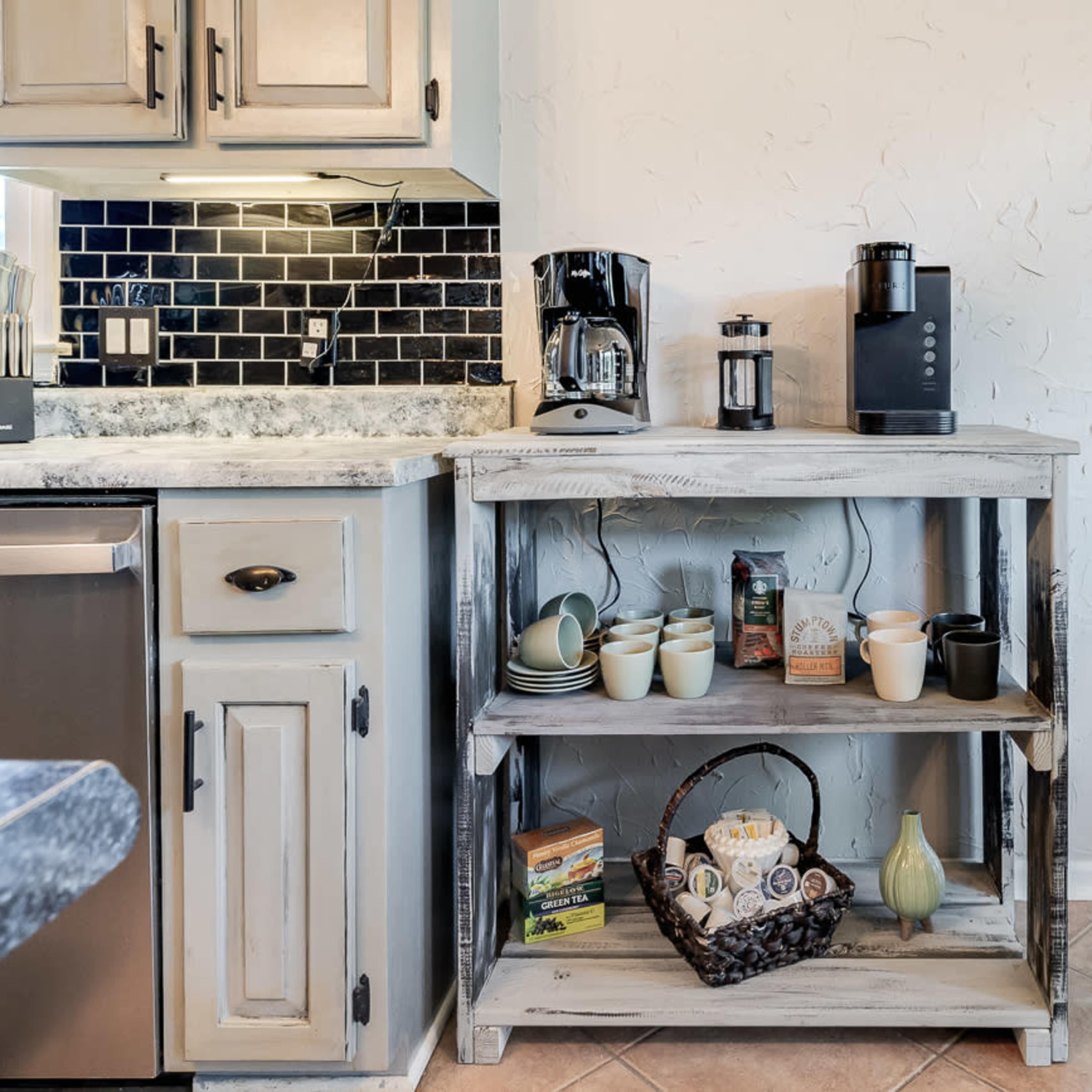 A wooden kitchen cart with two shelves displays various coffee-making appliances and mugs, situated beside a stainless steel refrigerator.
