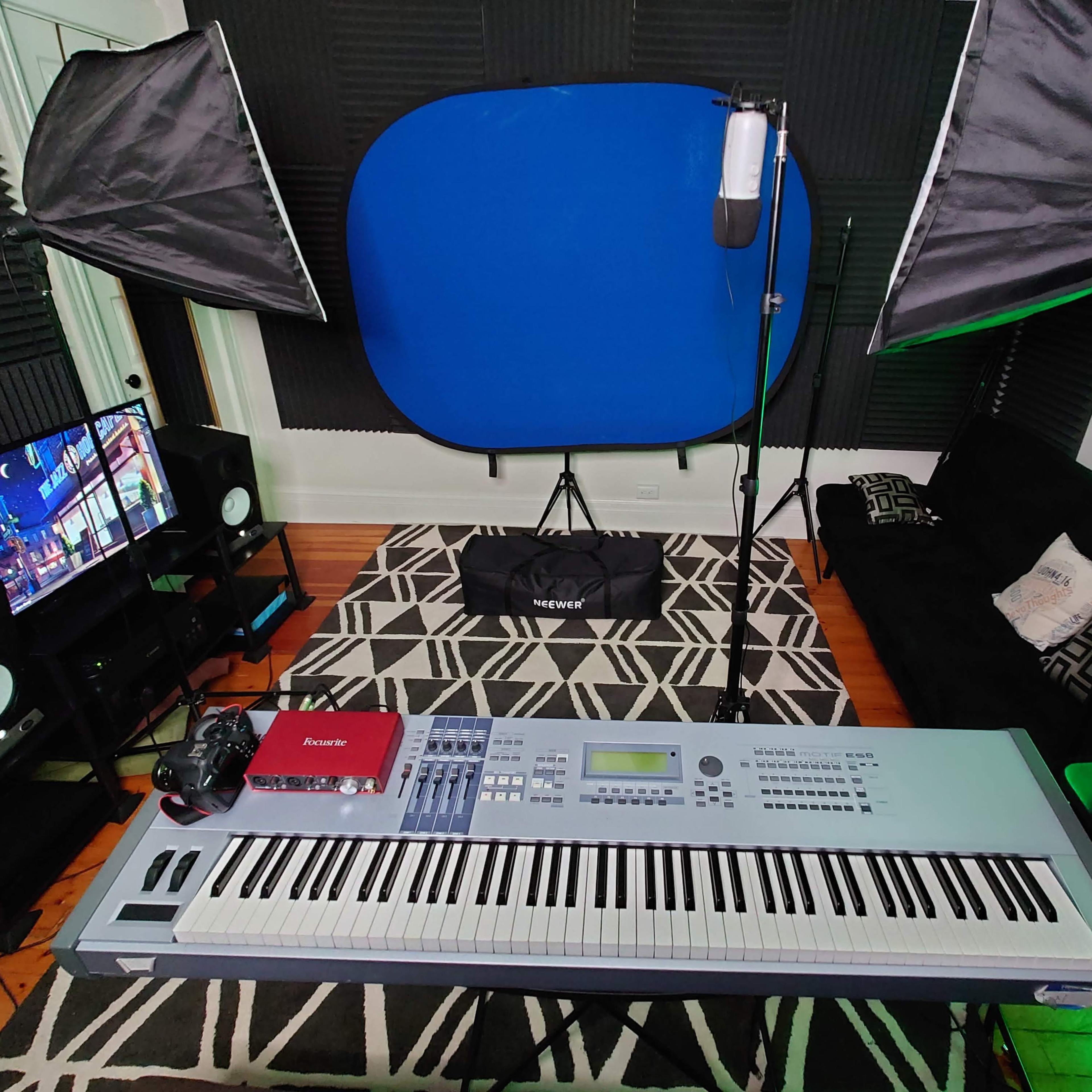 A music production setup with a keyboard, a microphone, lighting equipment, a monitor, and a laptop arranged in a room with acoustic panels and a blue backdrop.