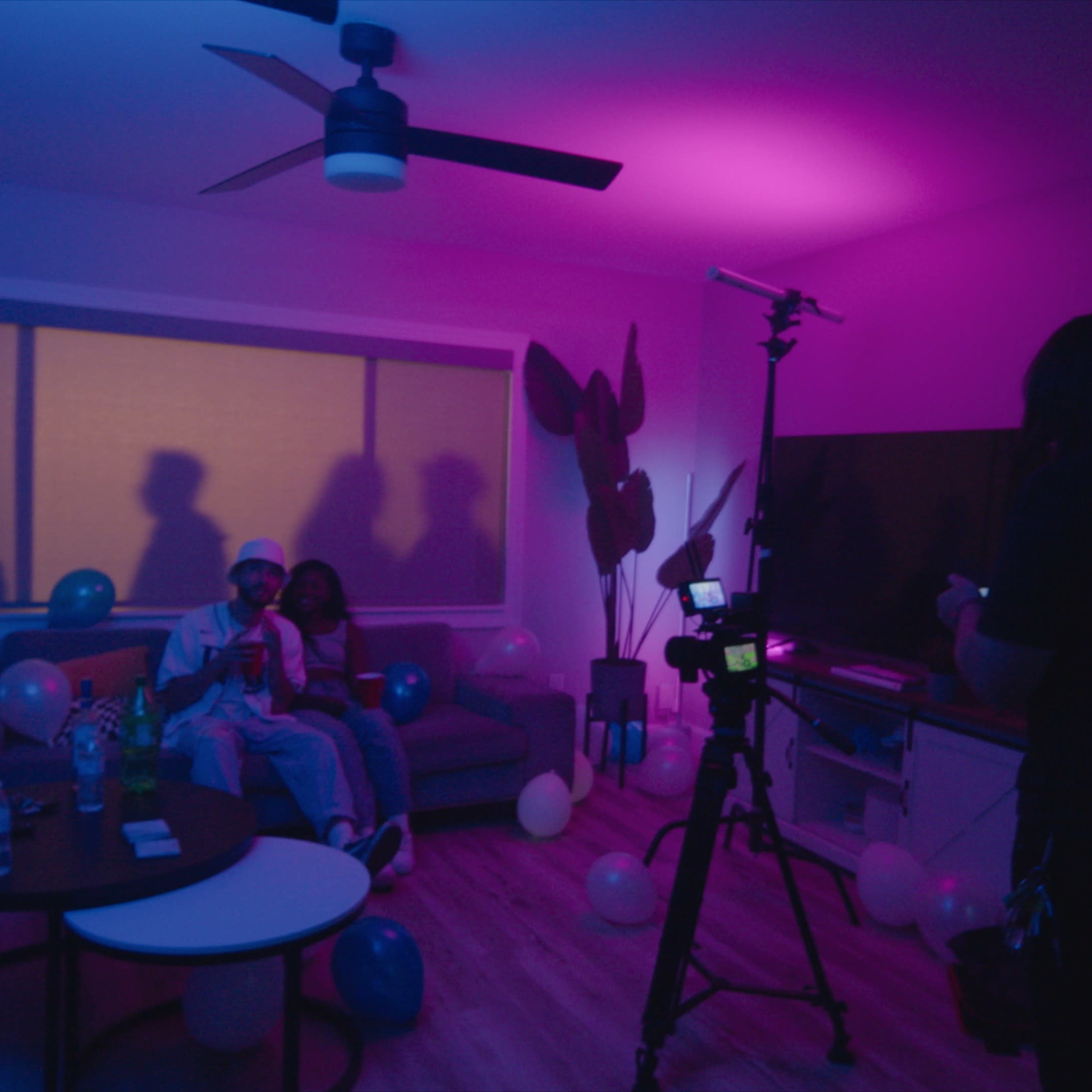A small group of people poses on a couch in a dimly lit room decorated with balloons, while a camera and lighting equipment are set up nearby.
