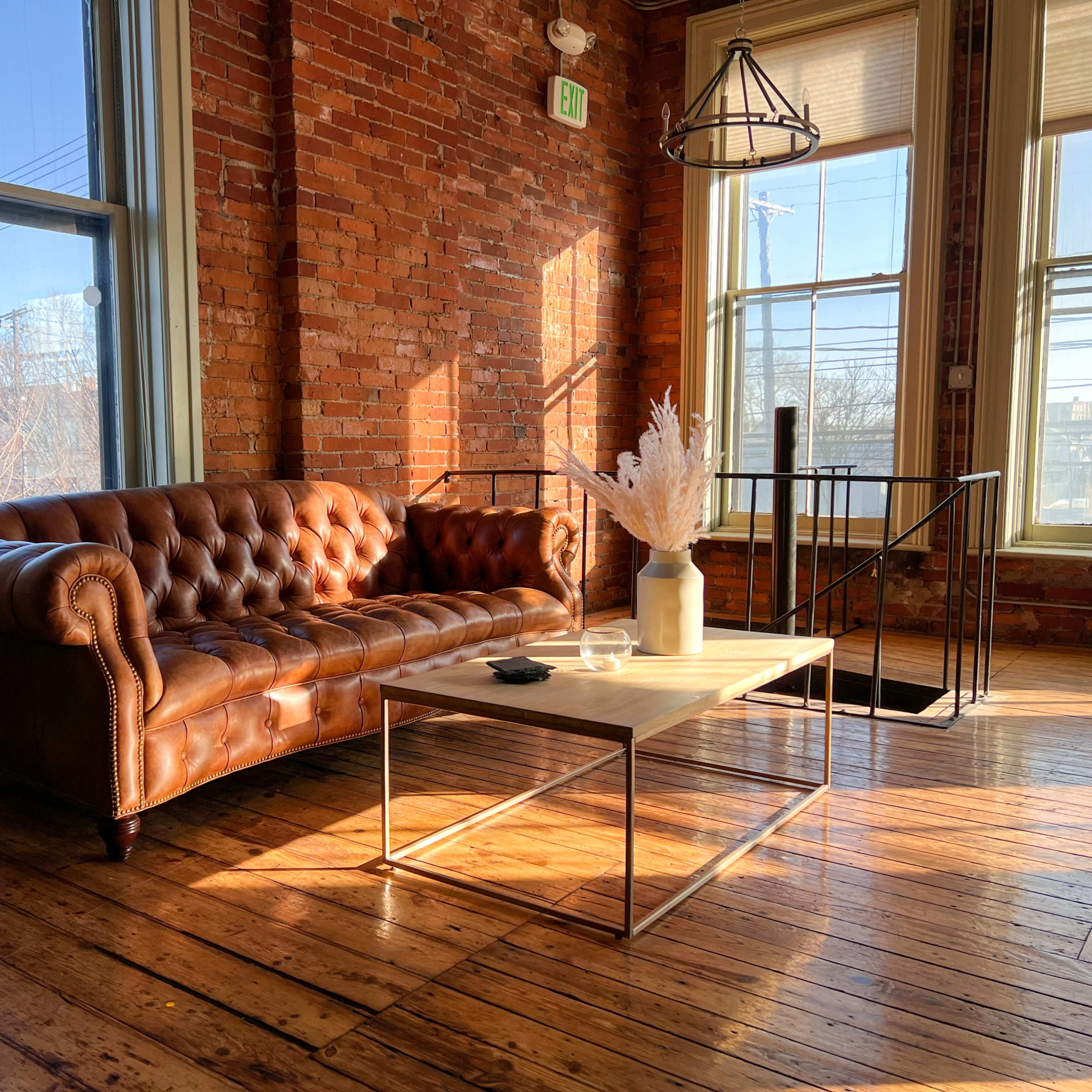 A leather chesterfield sofa and a wooden coffee table in a sunlit room with exposed brick walls and large windows.