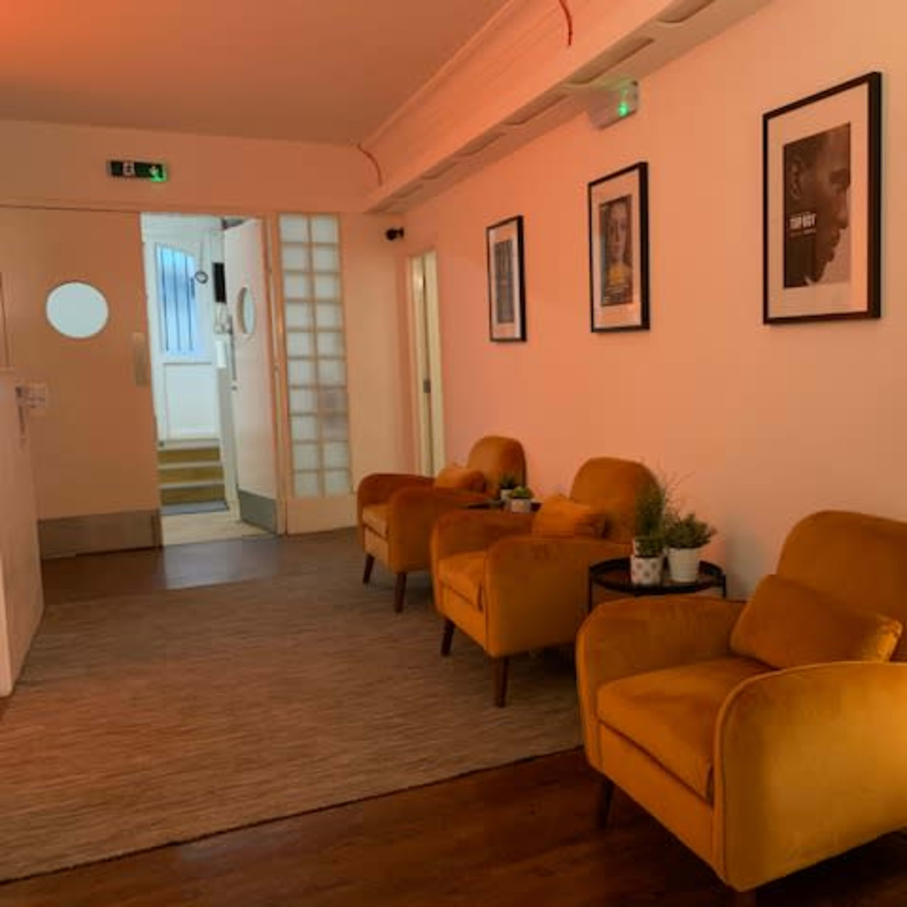 A hallway with three yellow armchairs, framed photographs on the walls, and a standing lamp, leading to a door at the end.