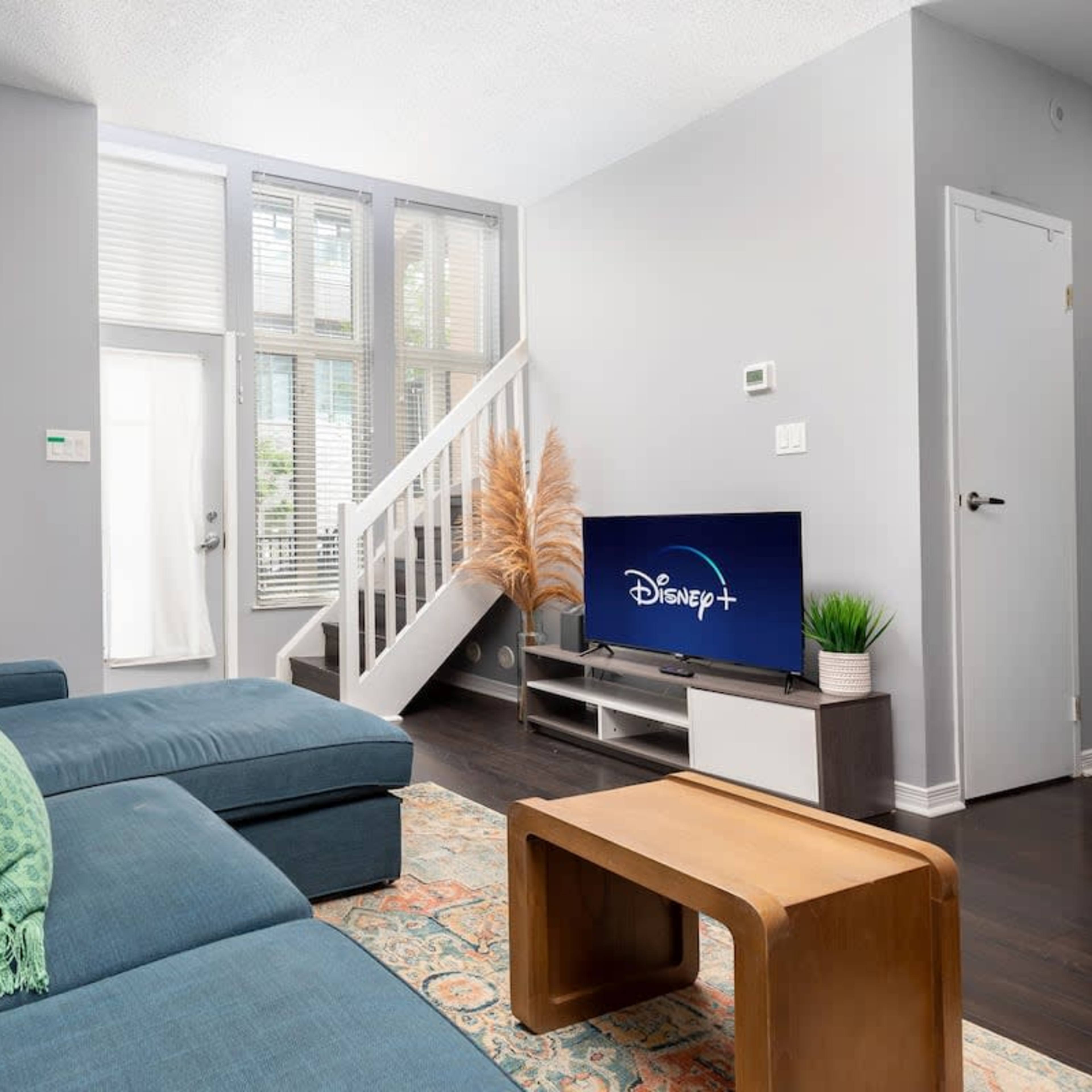 A living room with a blue sectional sofa, a wooden coffee table, and a television displaying the Disney+ logo, accompanied by large windows and a staircase in the background.