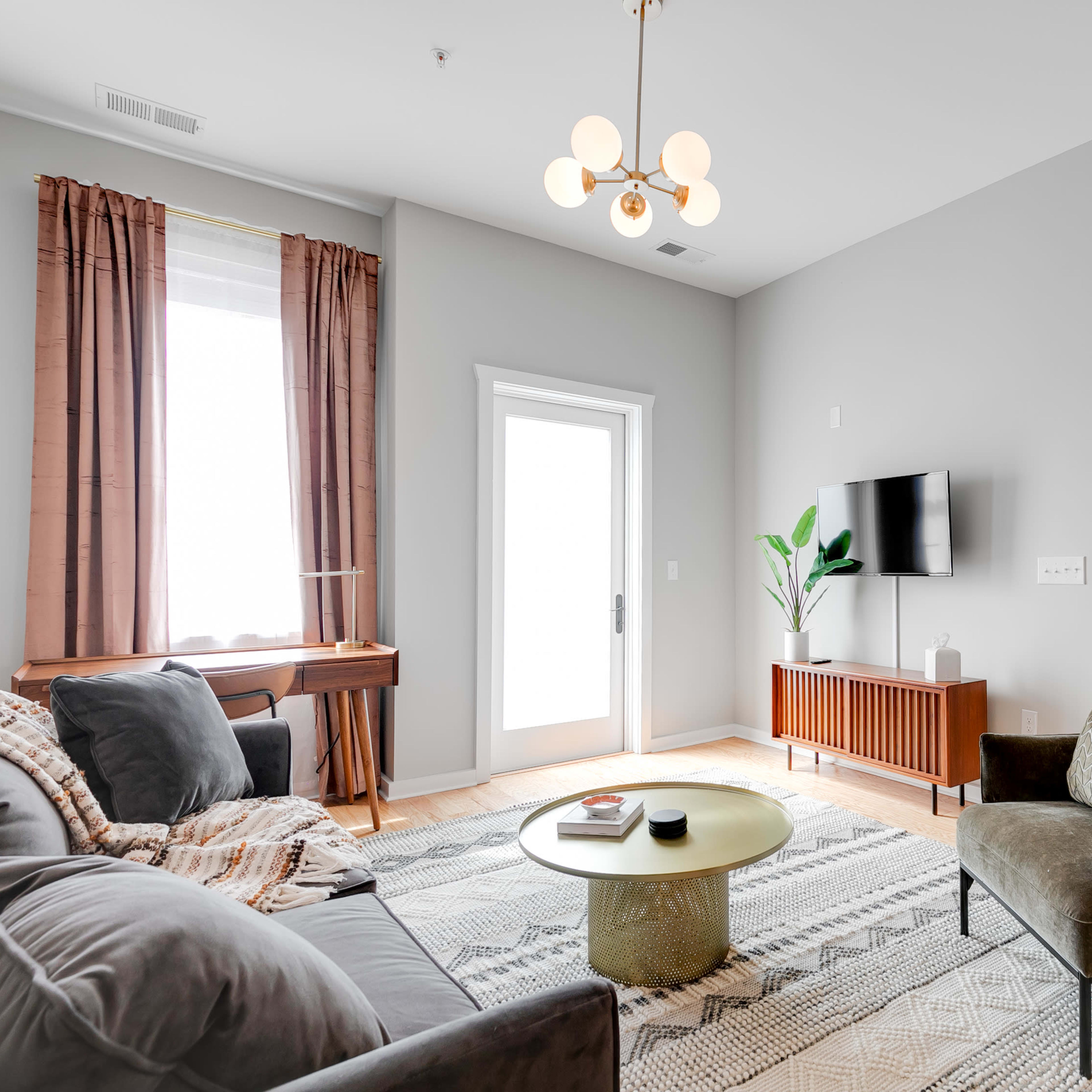 A modern living room features a gray sofa and armchair, a round coffee table, a television mounted on the wall, and large windows with curtains.