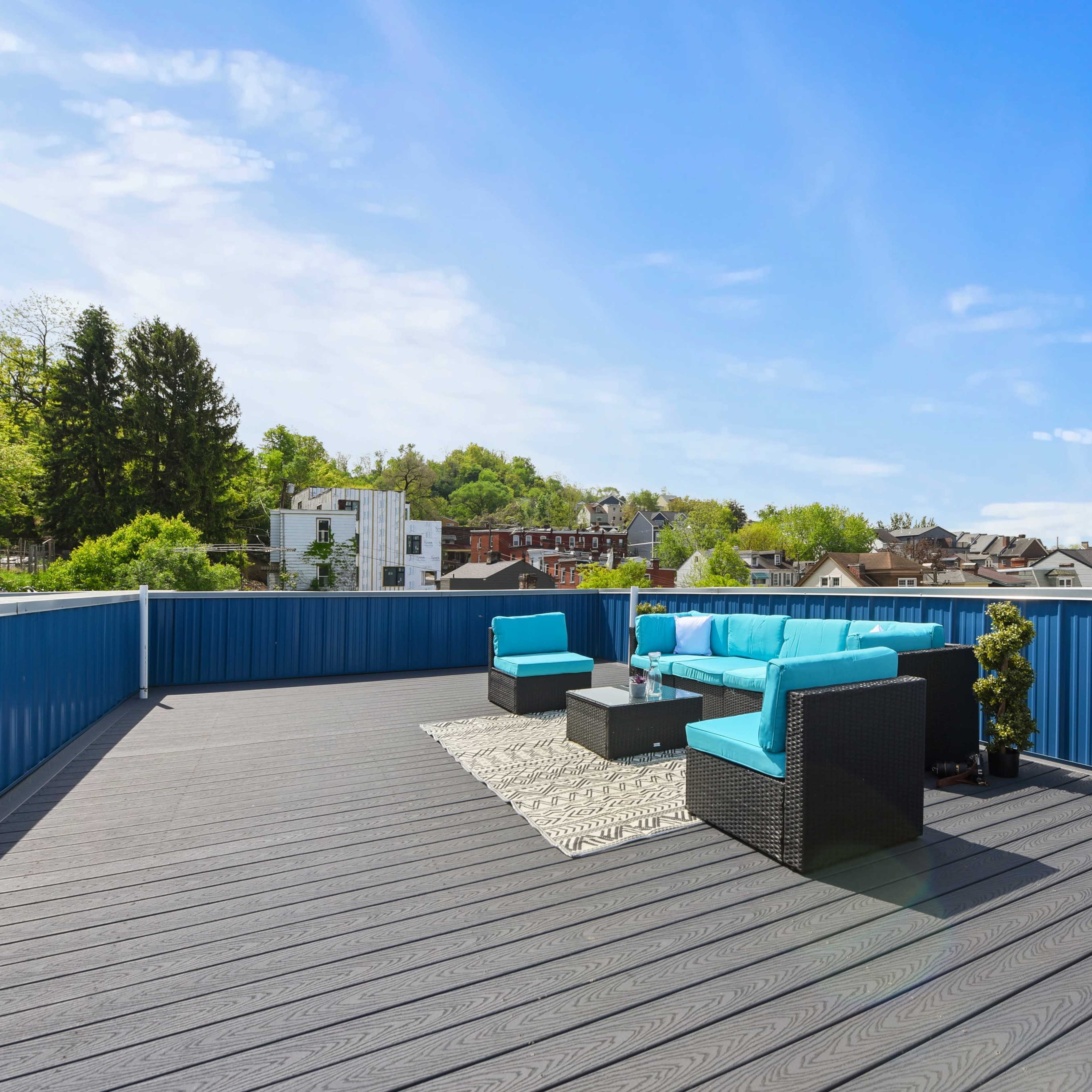 A rooftop terrace features a turquoise seating area surrounded by trees and buildings under a clear blue sky.