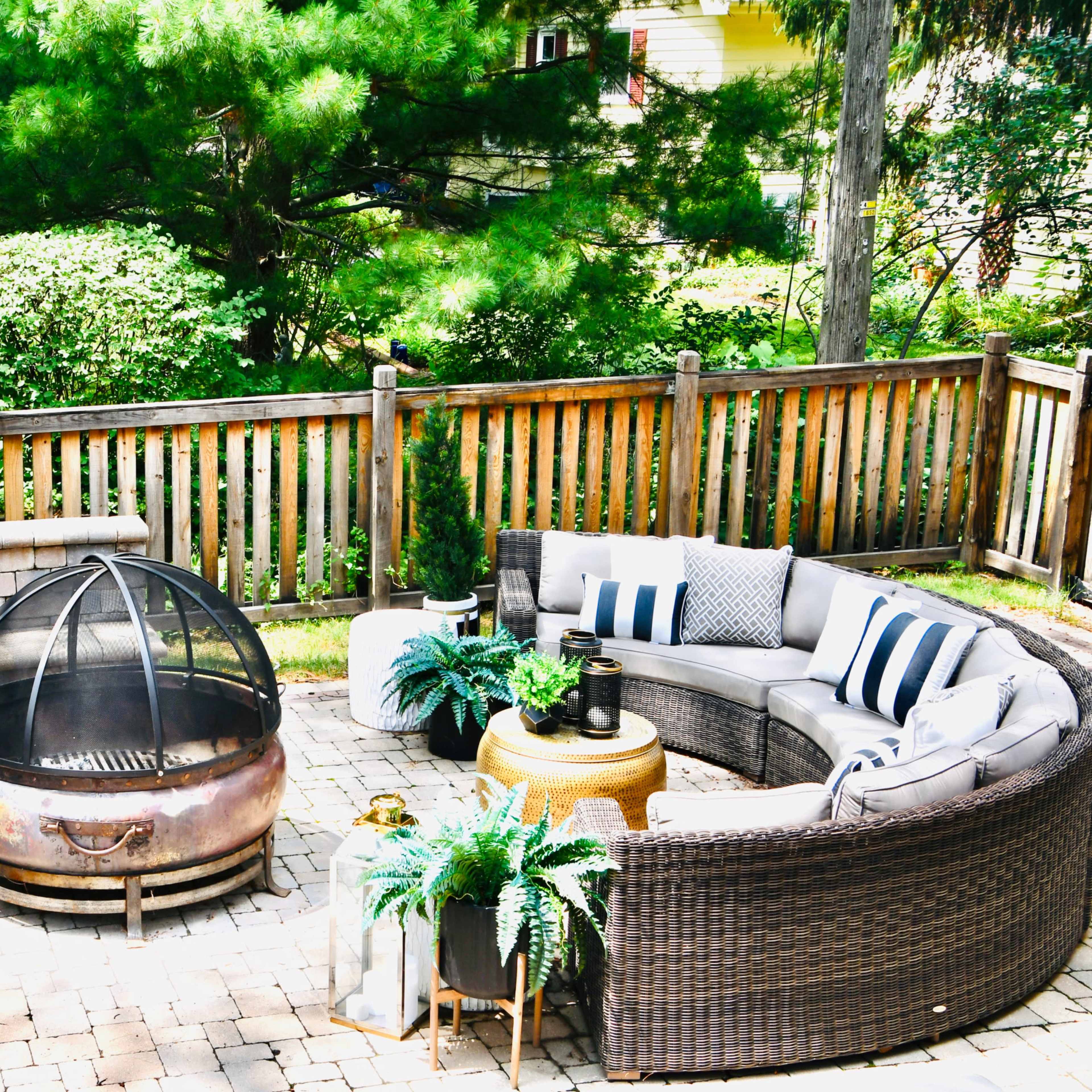 A landscaped backyard featuring a circular stone fire pit surrounded by a curved wicker sofa and decorative plants.