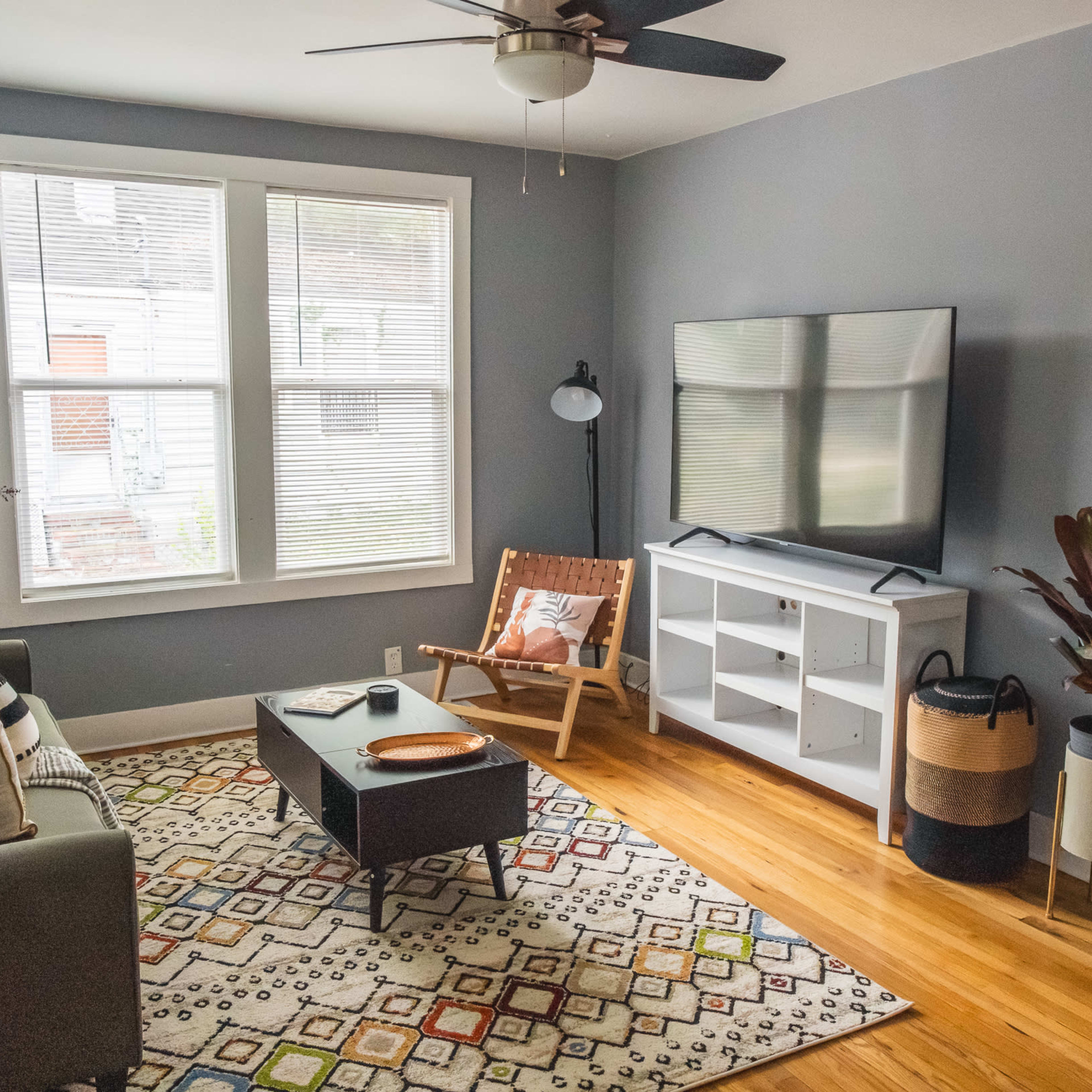 A cozy living room features a green sofa, a coffee table, a television on a white stand, and large windows with natural light.
