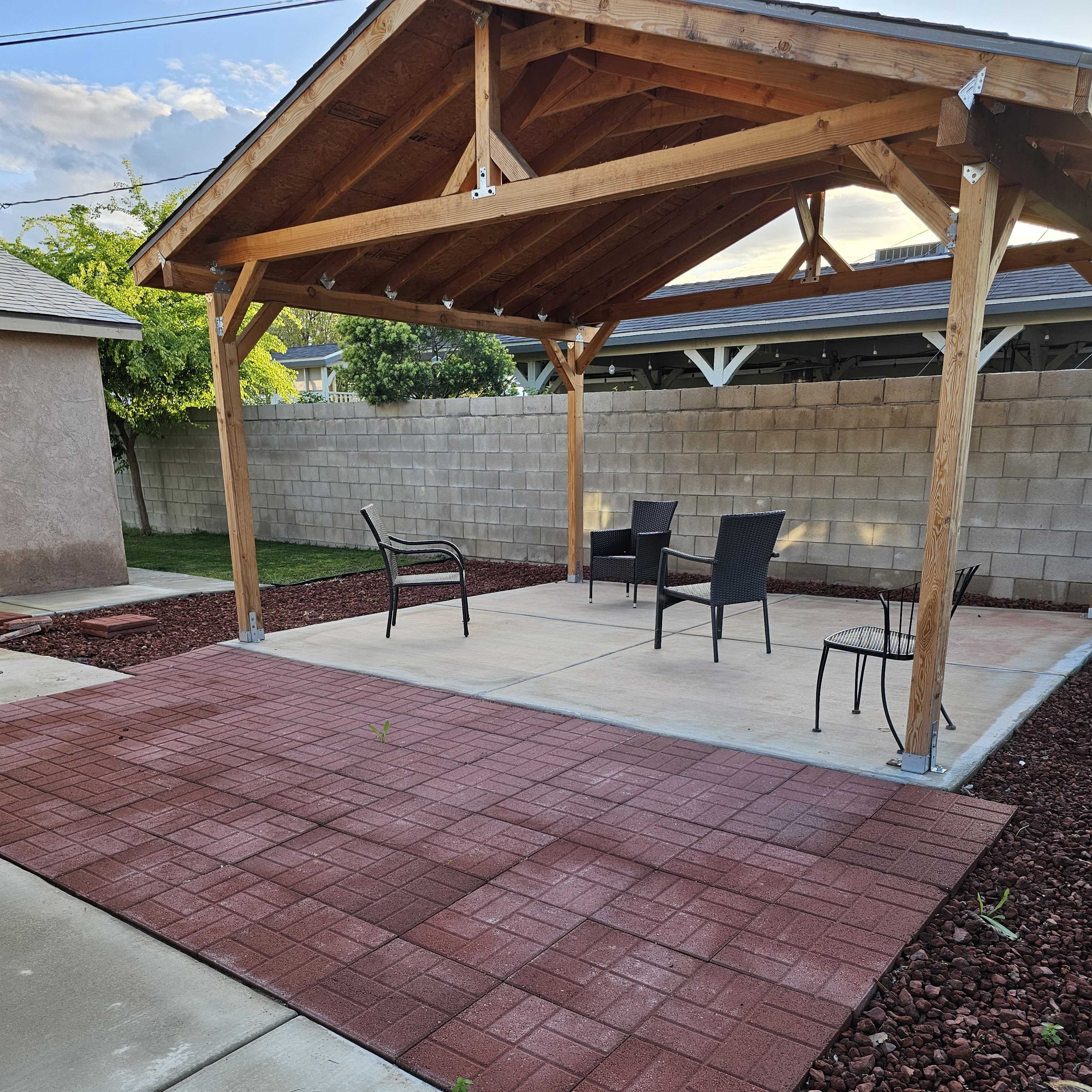 A patio area with a wooden pavilion, surrounded by gravel and featuring several chairs on a concrete surface.