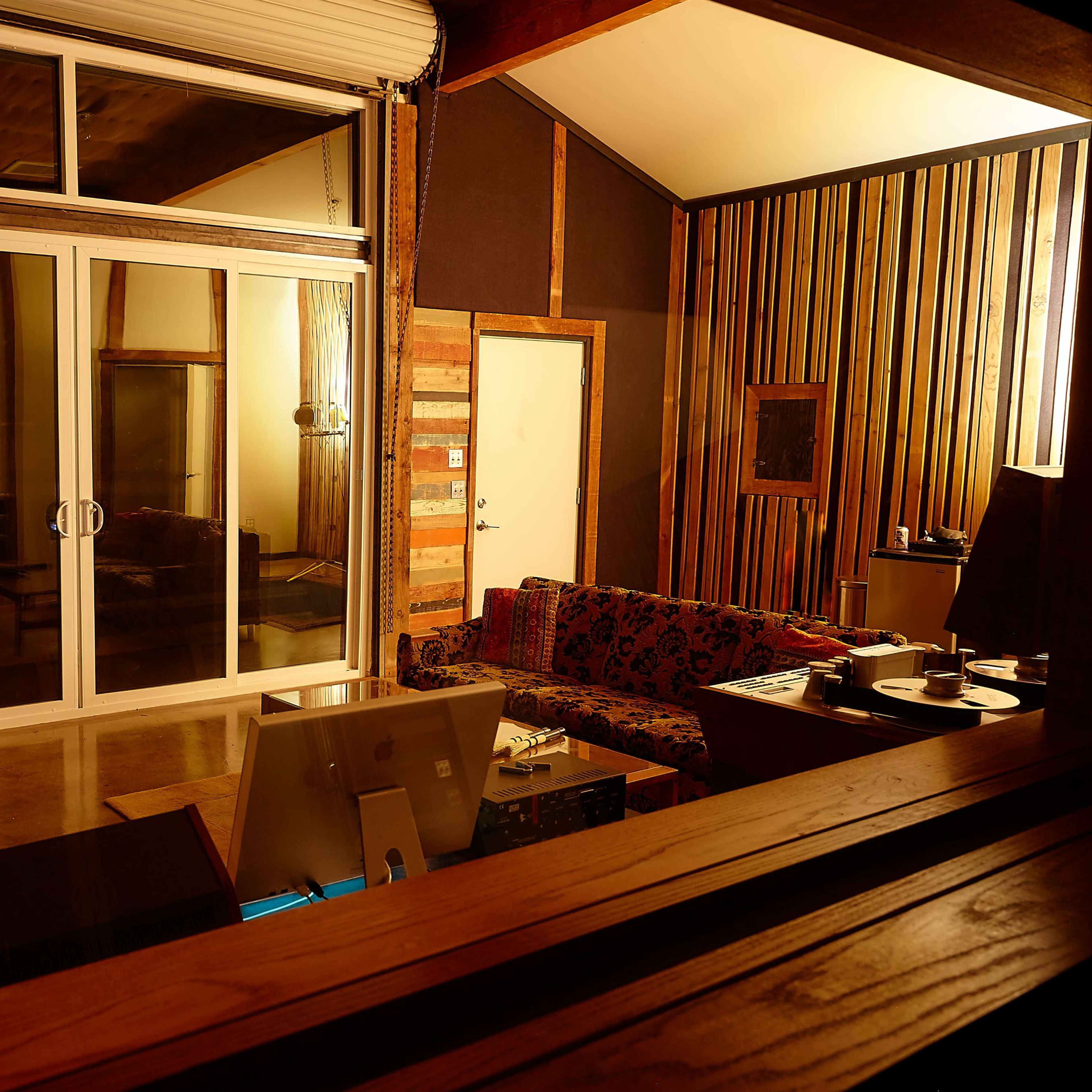 A modern recording studio with a mixing console, a computer workstation, a colorful couch, and large glass doors leading to an outdoor area.