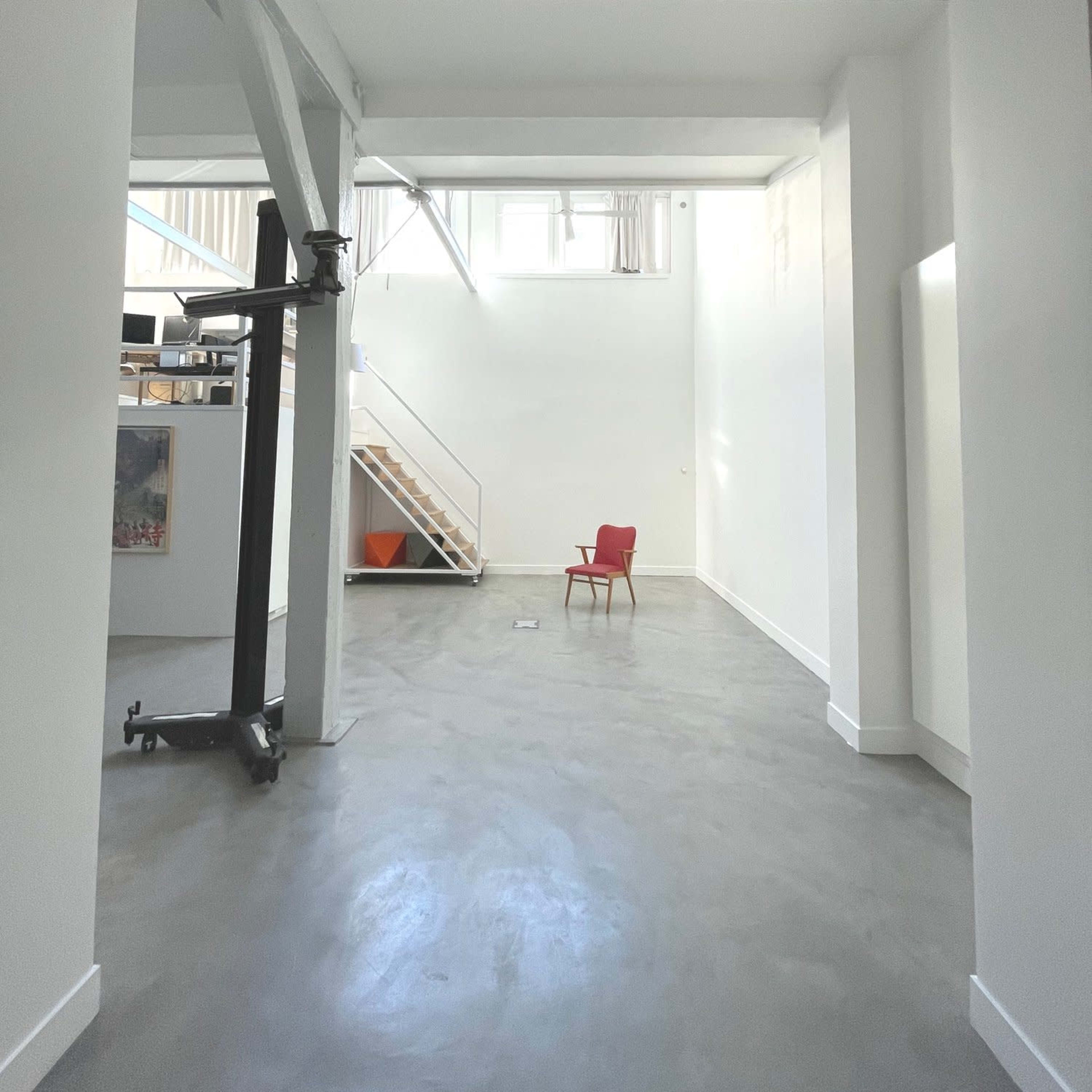 A spacious, minimalist interior with high white walls, exposed beams, and a single red chair positioned in the center.