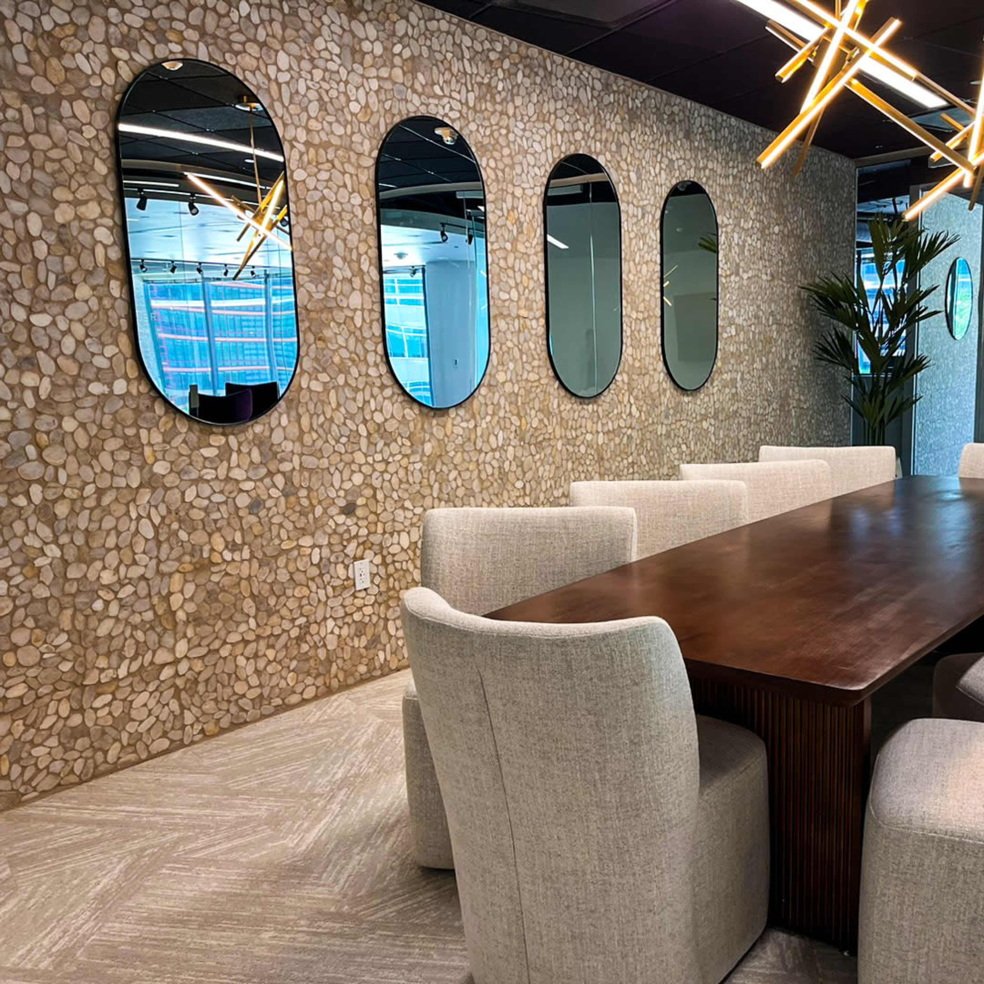 A long wooden table is surrounded by upholstered chairs in a modern conference room featuring a stone accent wall and decorative mirrors.
