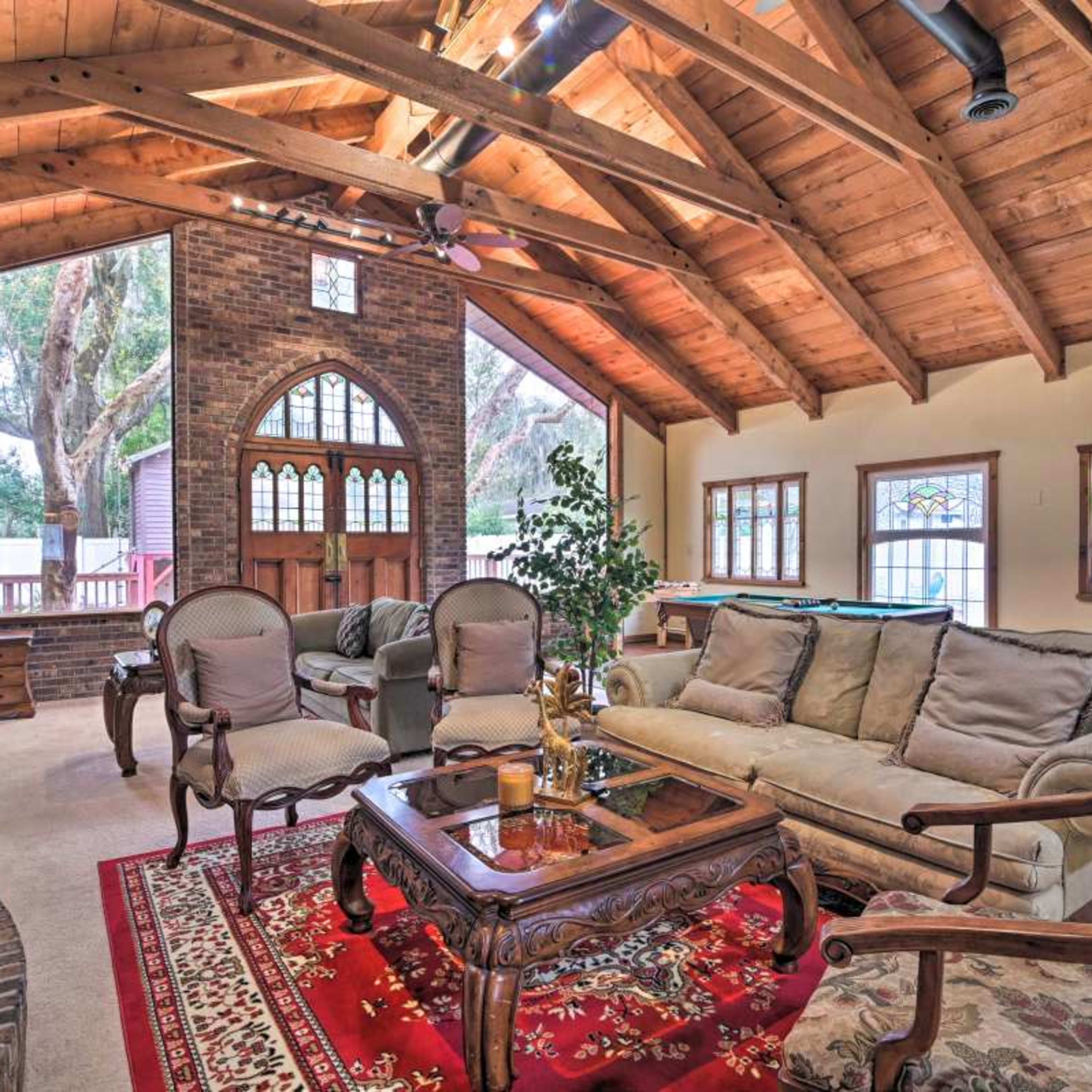 A spacious living room with a wooden ceiling, a brick fireplace, and a mix of furniture including a sofa, armchairs, and a coffee table, all arranged around a patterned rug.