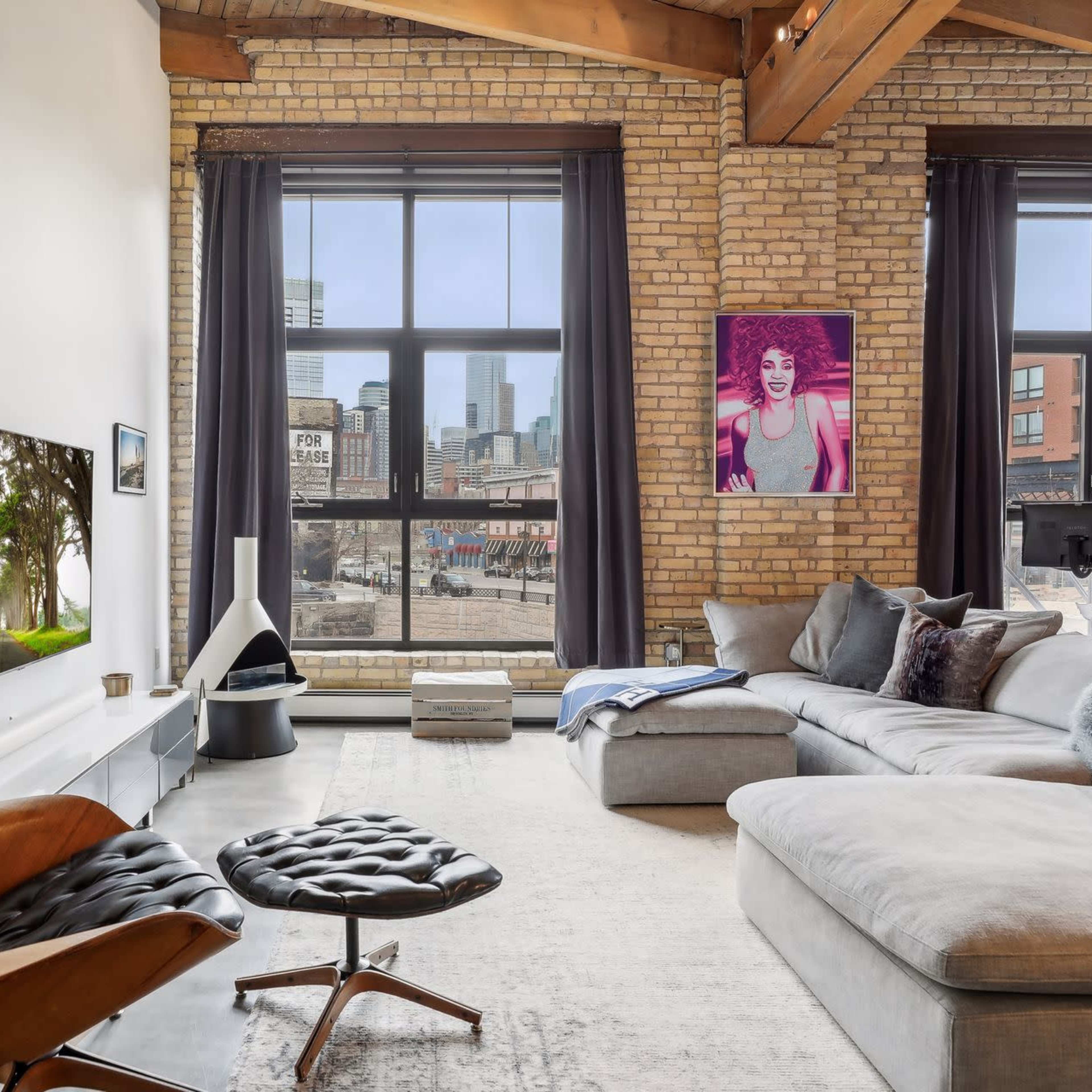 A modern living room features a sectional sofa, a sleek TV mounted on a brick wall, and large windows showcasing a city view.