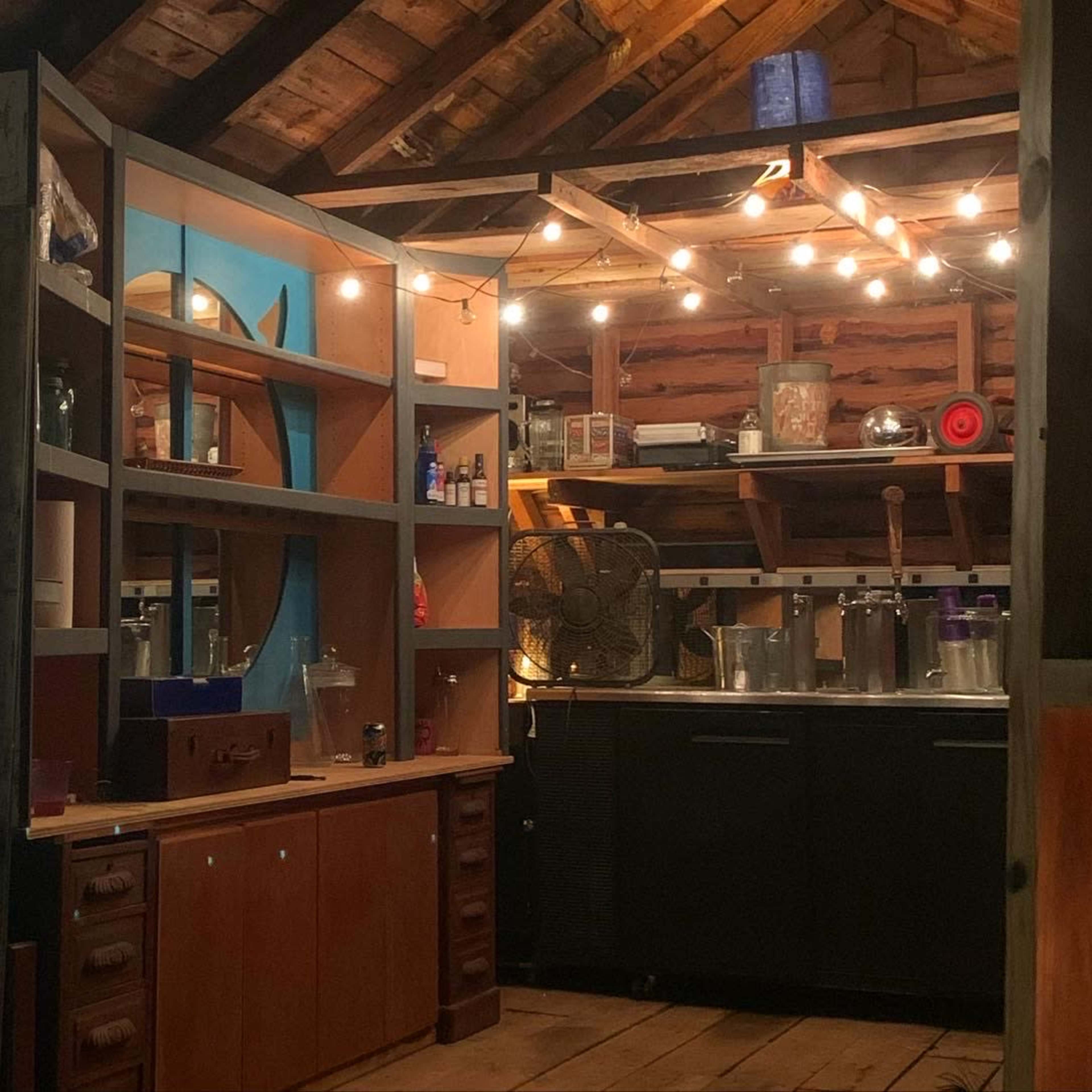 A rustic kitchen area in an attic, featuring wooden cabinetry, shelves with various items, and string lights illuminating the space.