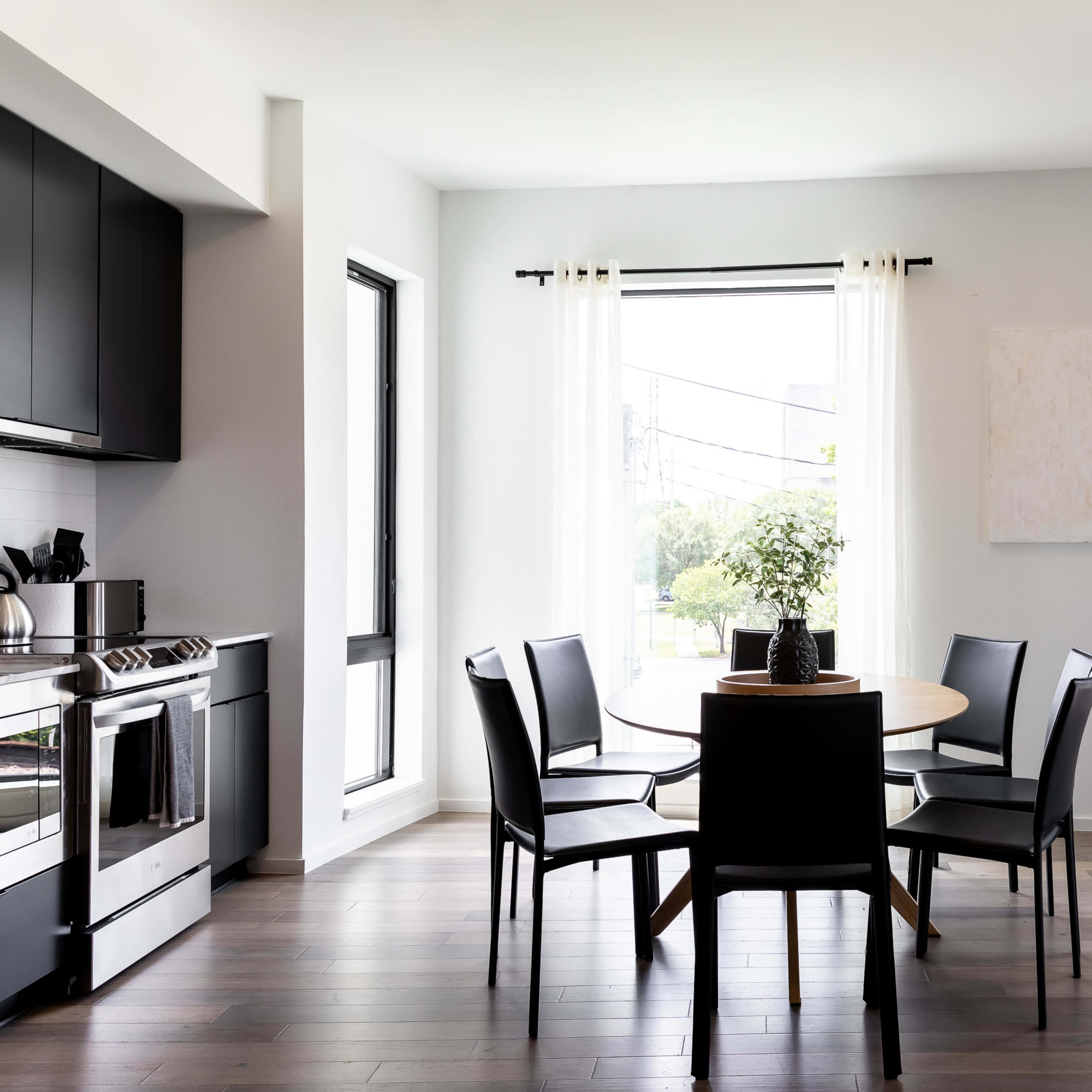A dining area features a round table surrounded by four black chairs, with large windows letting in natural light.