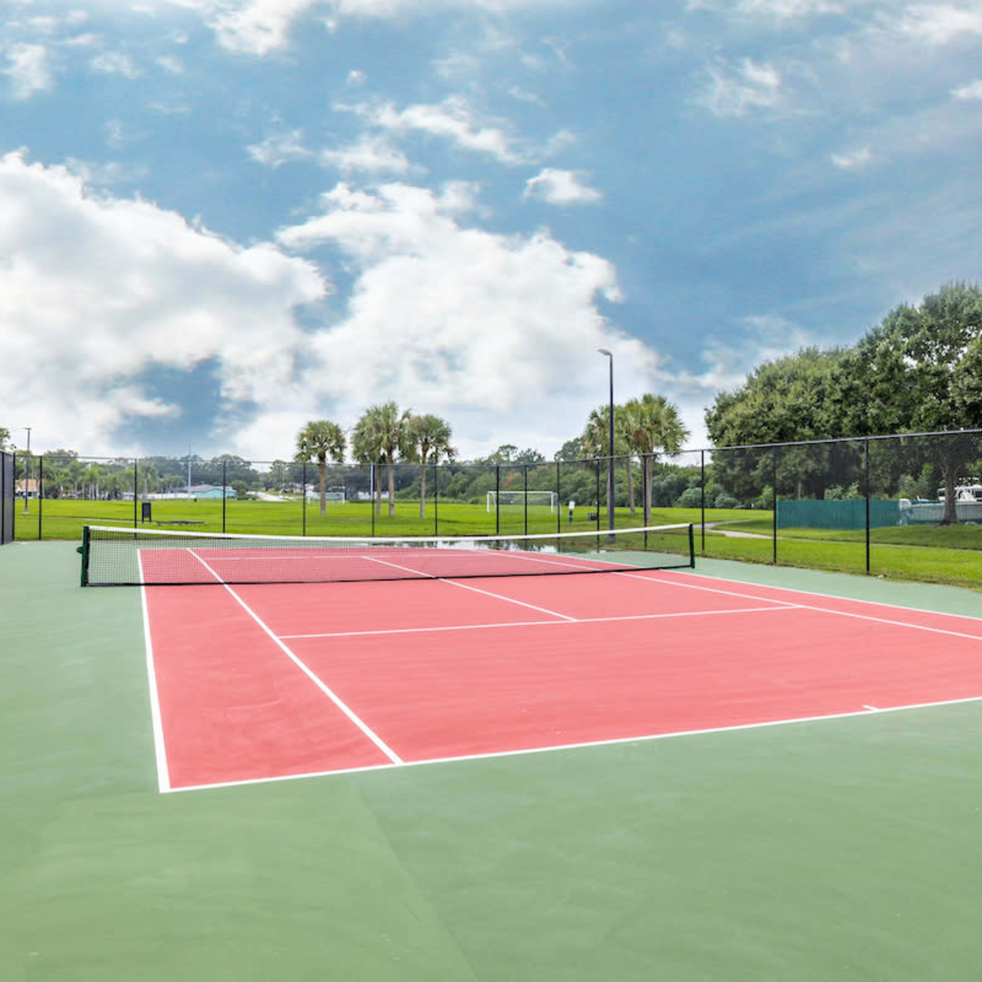 A well-maintained tennis court with red and green surfaces under a partly cloudy sky.