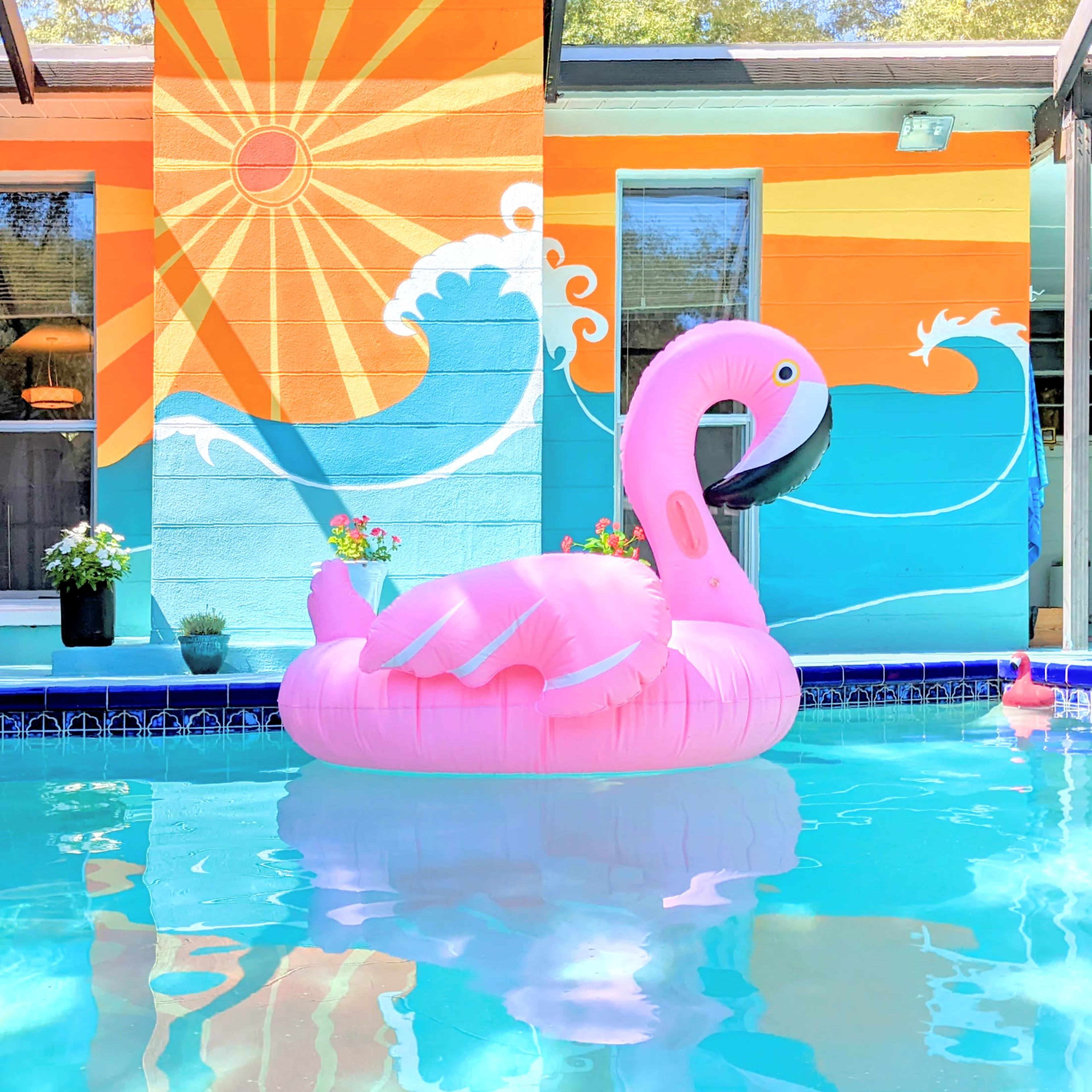 A large pink flamingo float sits in a blue swimming pool with a colorful mural of waves and a sun on the wall behind it.