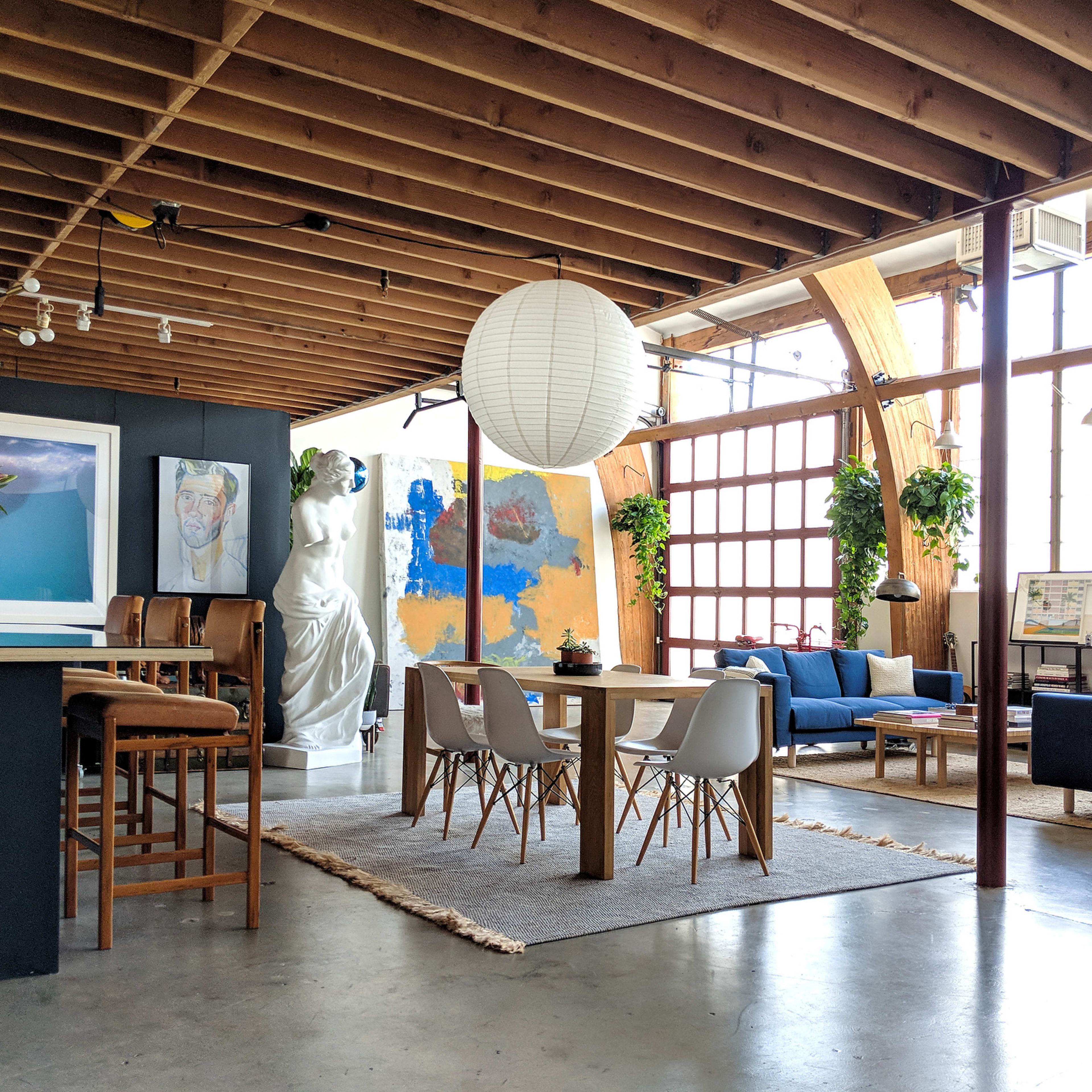 A stylish open-concept living space featuring a dining area with a large table, a kitchen bar, a sculpture, and various plants around the room.