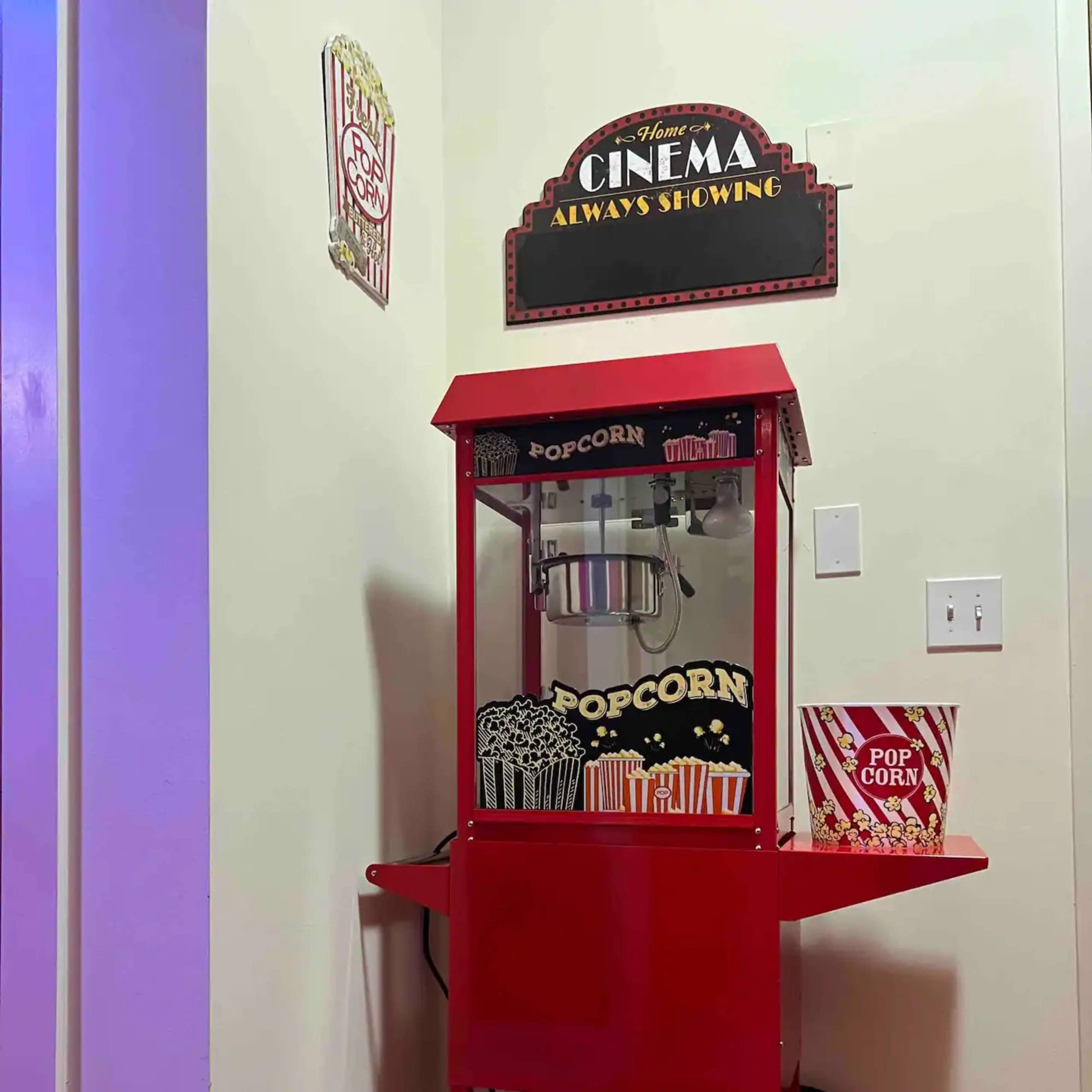 A red popcorn machine with a glass top sits against a wall, accompanied by a "Cinema Always Showing" sign and popcorn-themed decorations.