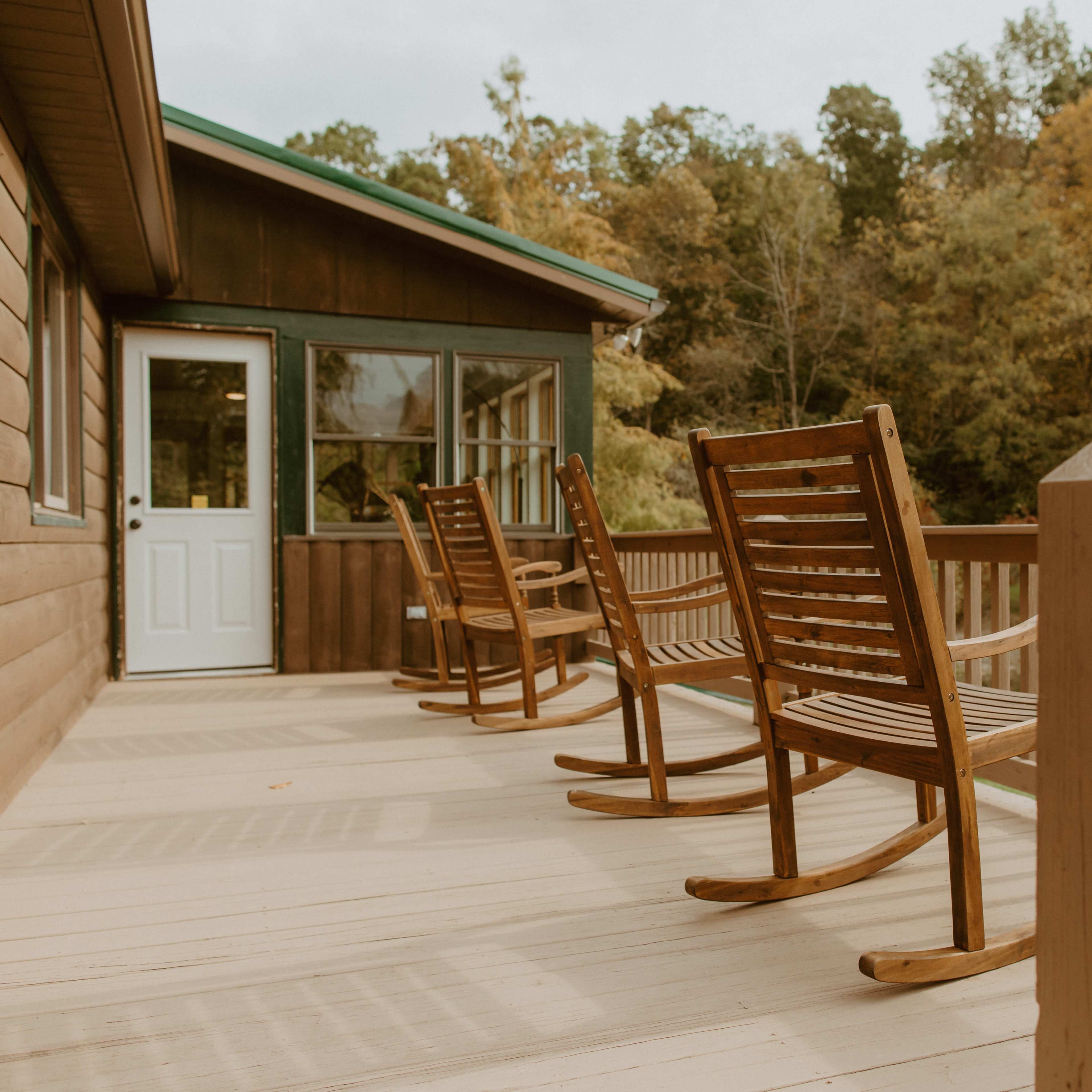 A wooden deck features three rocking chairs facing a wooded area.