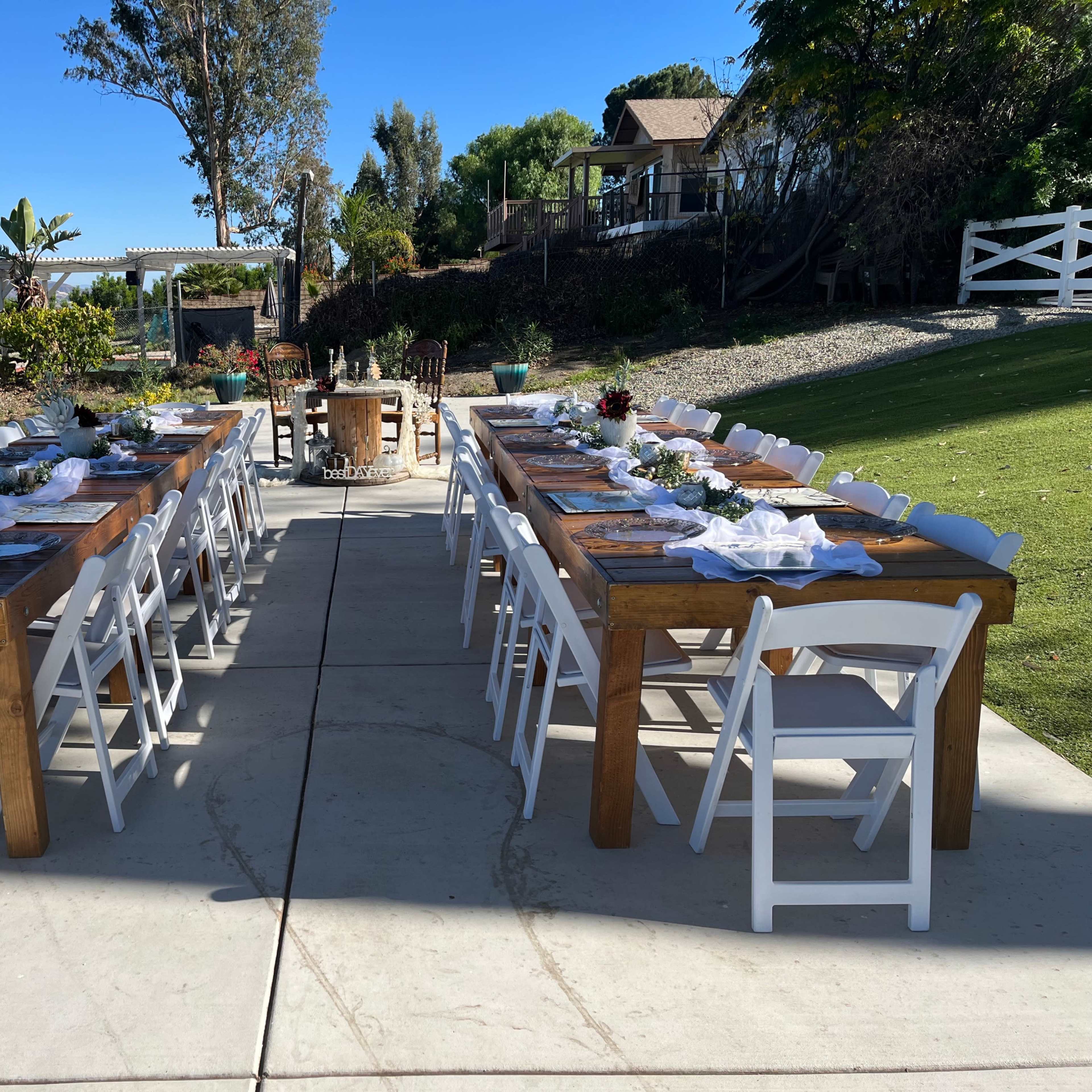 Two long wooden tables are set with white tableware and centerpieces, positioned on a paved area with greenery and a house in the background.