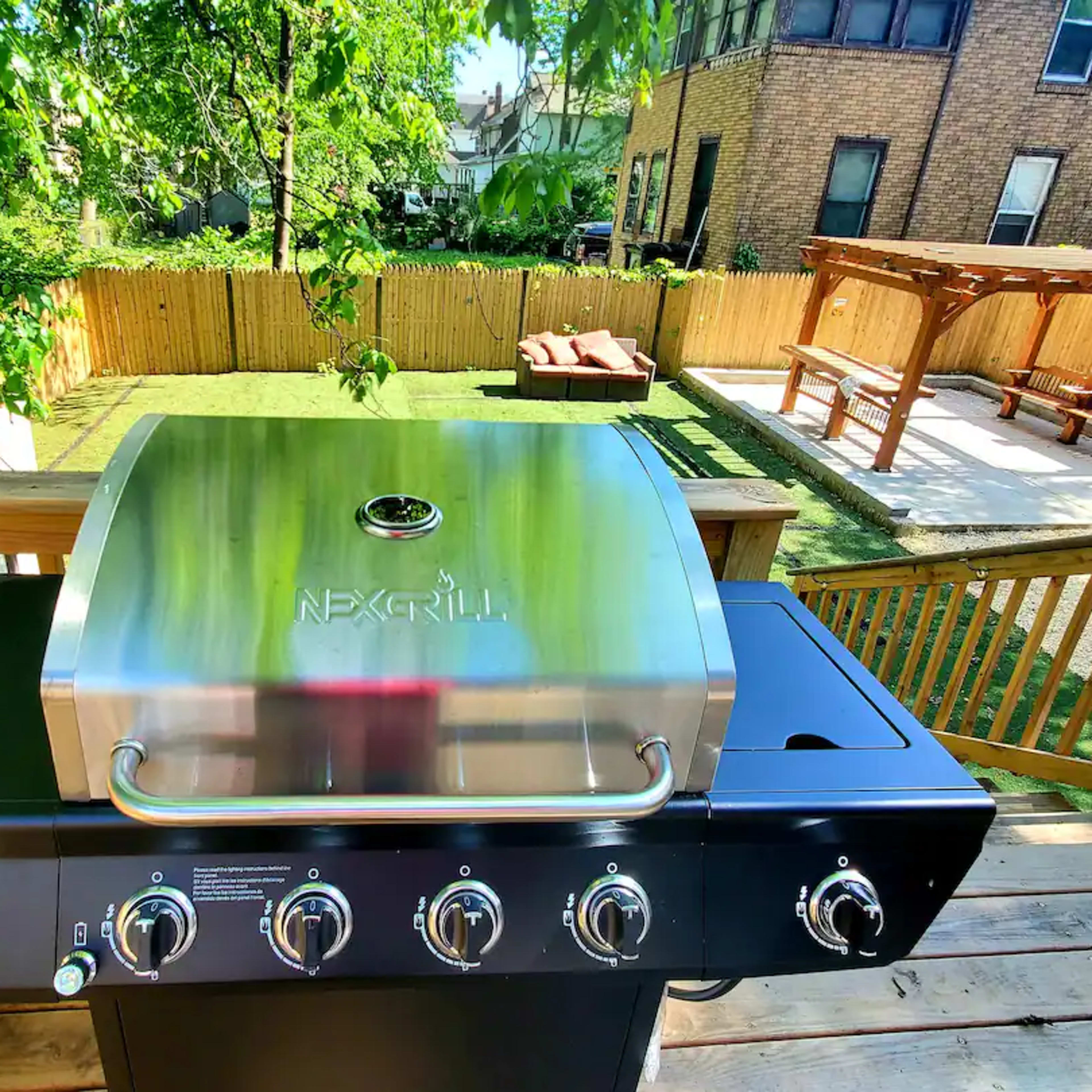 A stainless steel gas grill sits on a wooden deck, overlooking a backyard with a grassy area and a pergola.