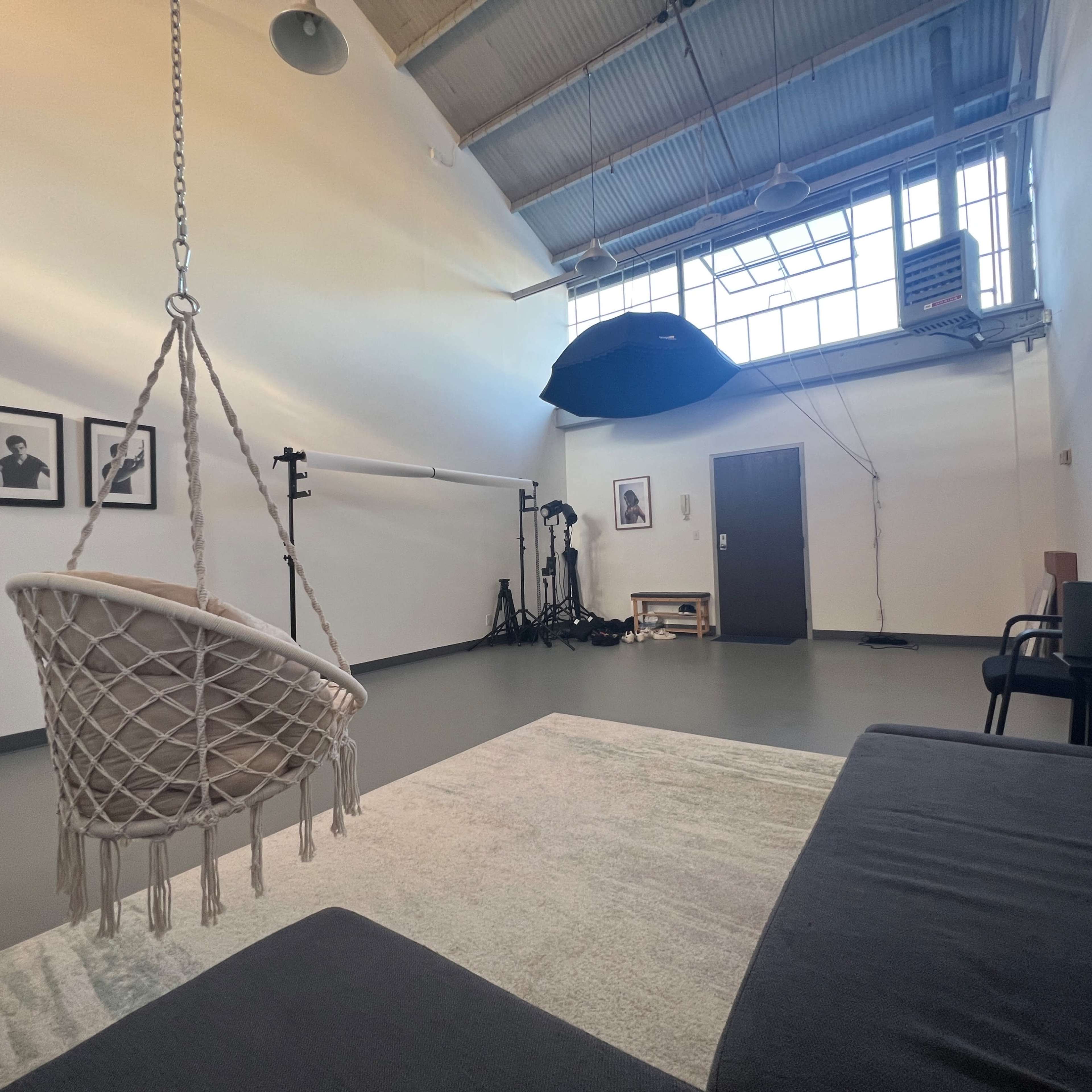 A spacious studio with a hanging chair, a sofa, framed photographs on the wall, and photography equipment in the background.