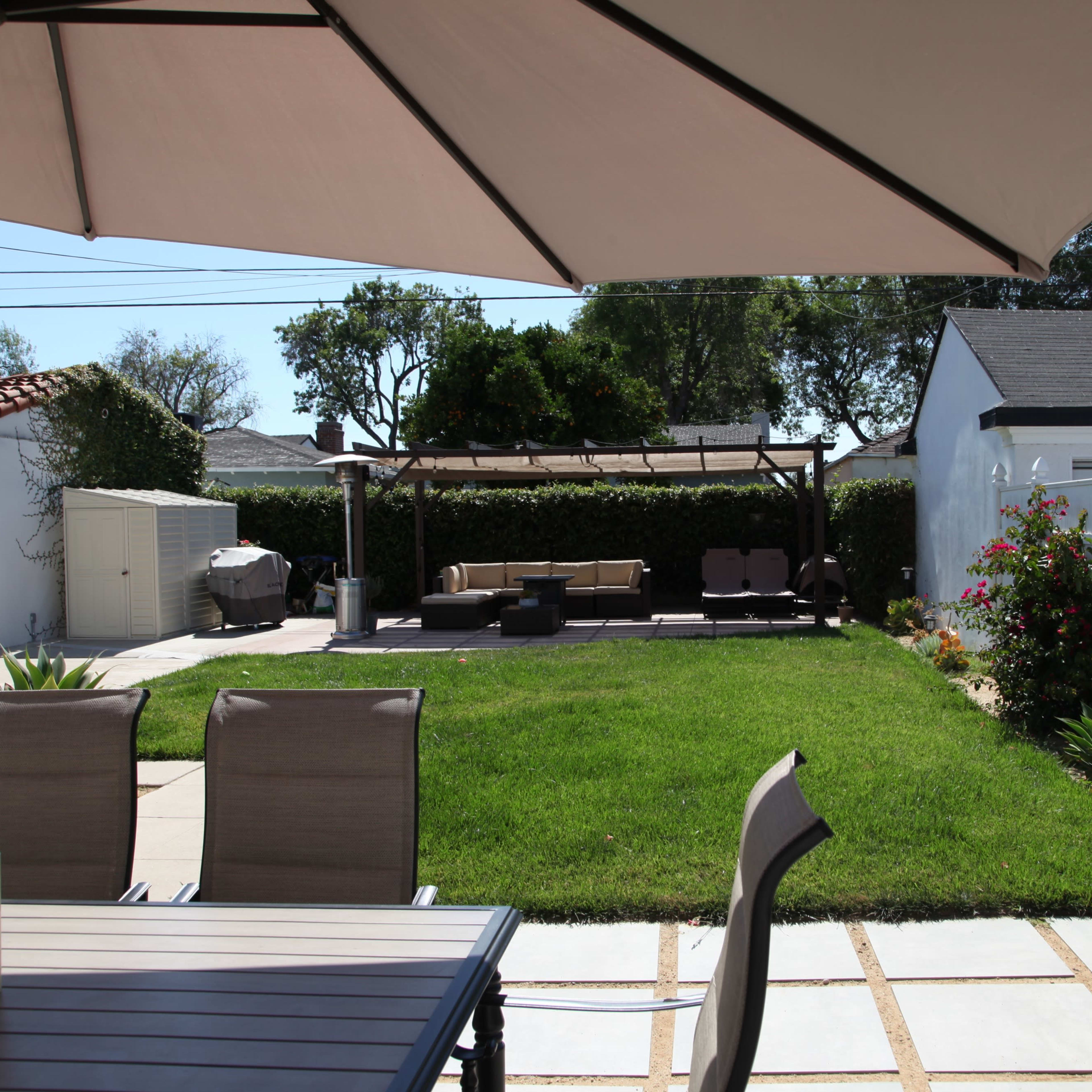 A patio area features a table and chairs in the foreground, with a grassy yard and seating area in the background under a large shade umbrella.