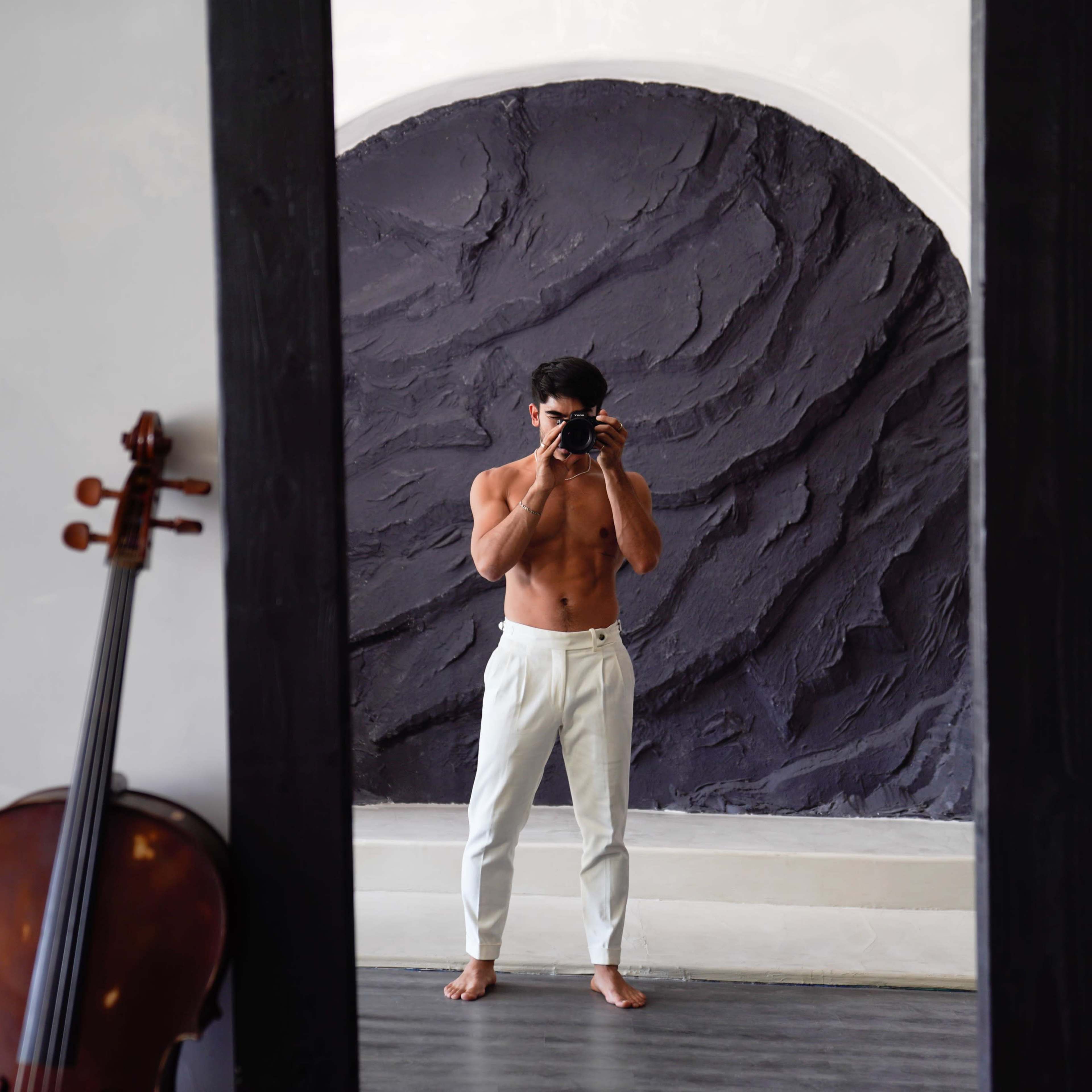 A shirtless man stands barefoot in front of a large mirror, holding a camera, with a cello leaning against the wall nearby.