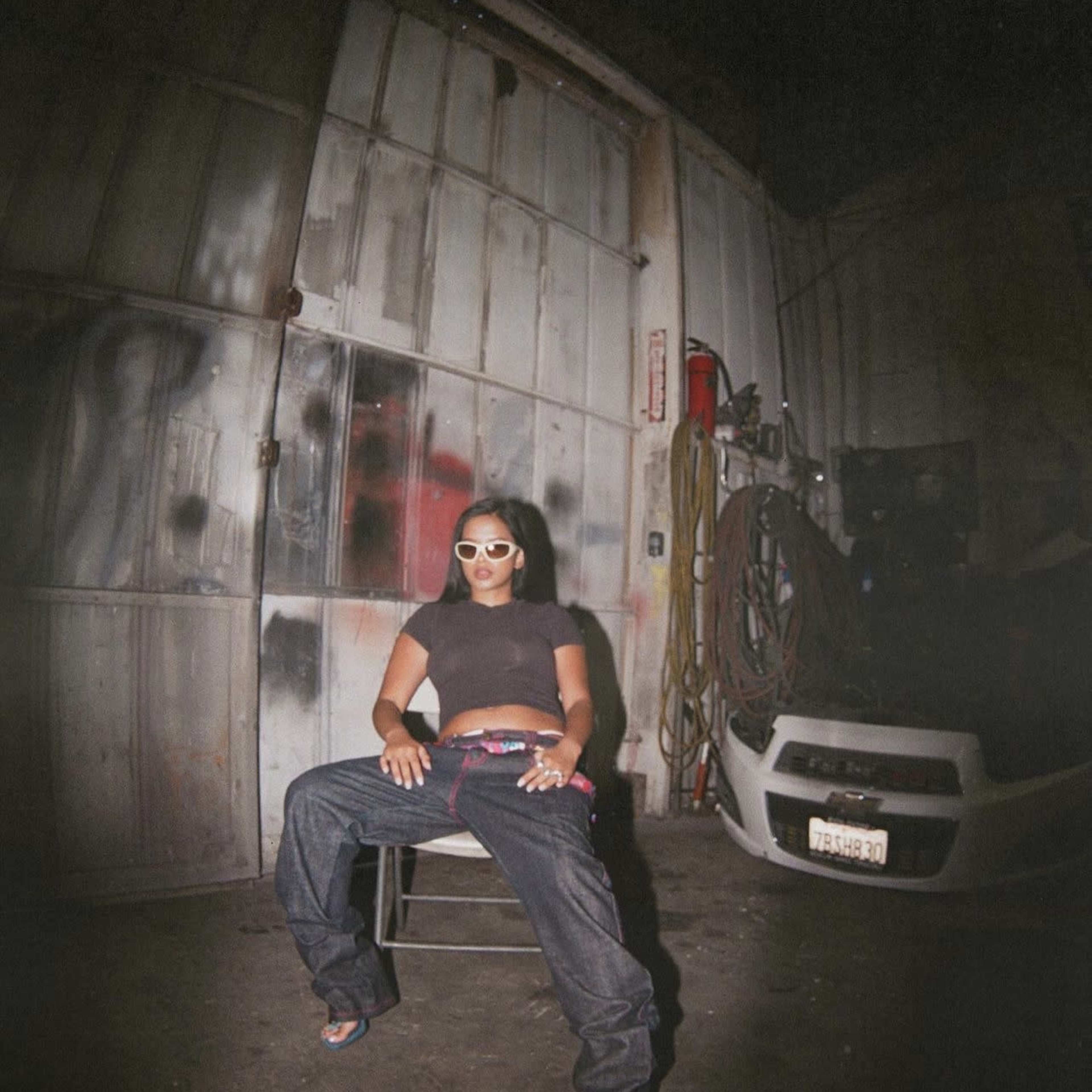 A woman wearing a black top and oversized jeans sits on a chair in a dimly lit industrial space.