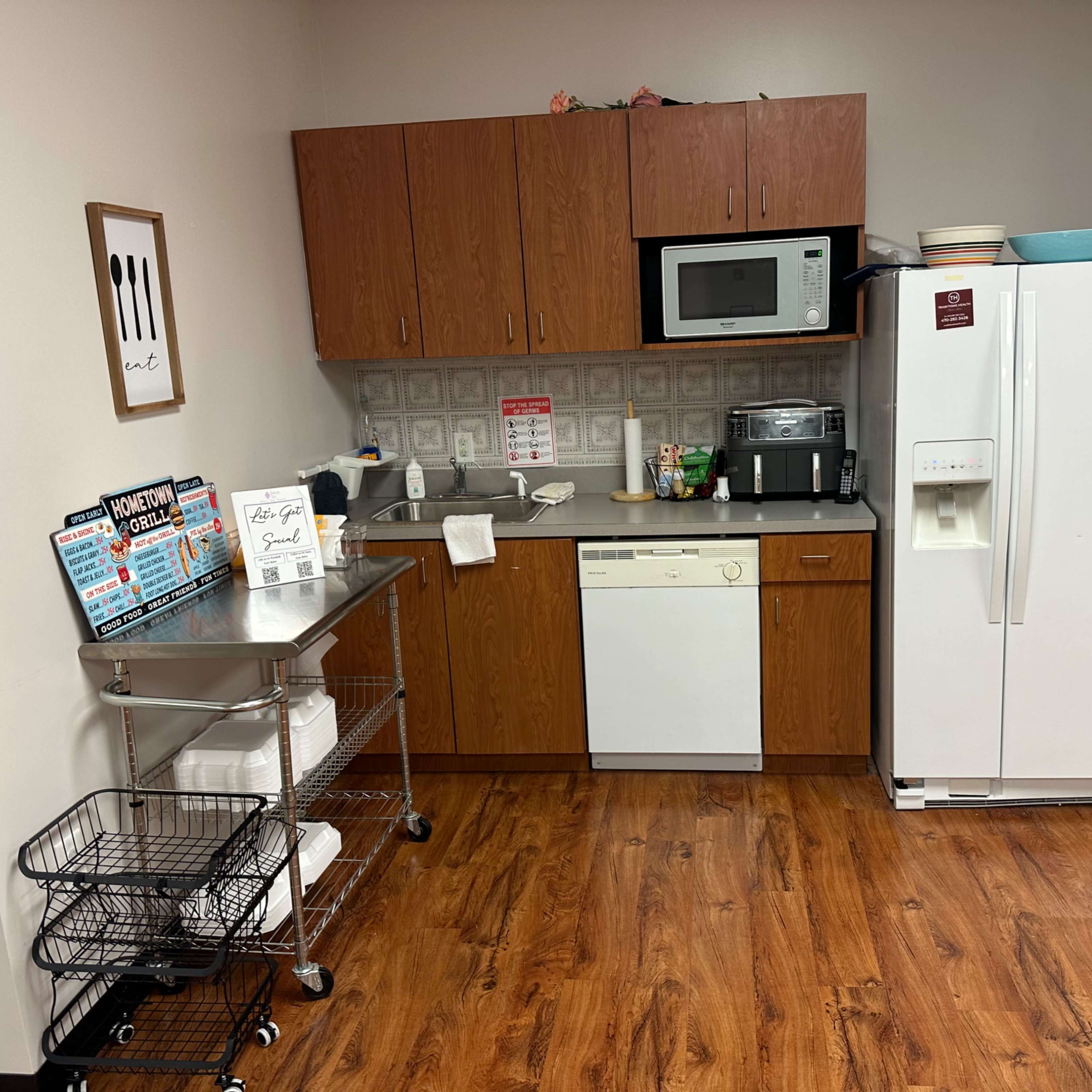 A kitchenette featuring a refrigerator, microwave, and dishwasher, along with wooden cabinets and a metal cart in a room with wooden flooring.