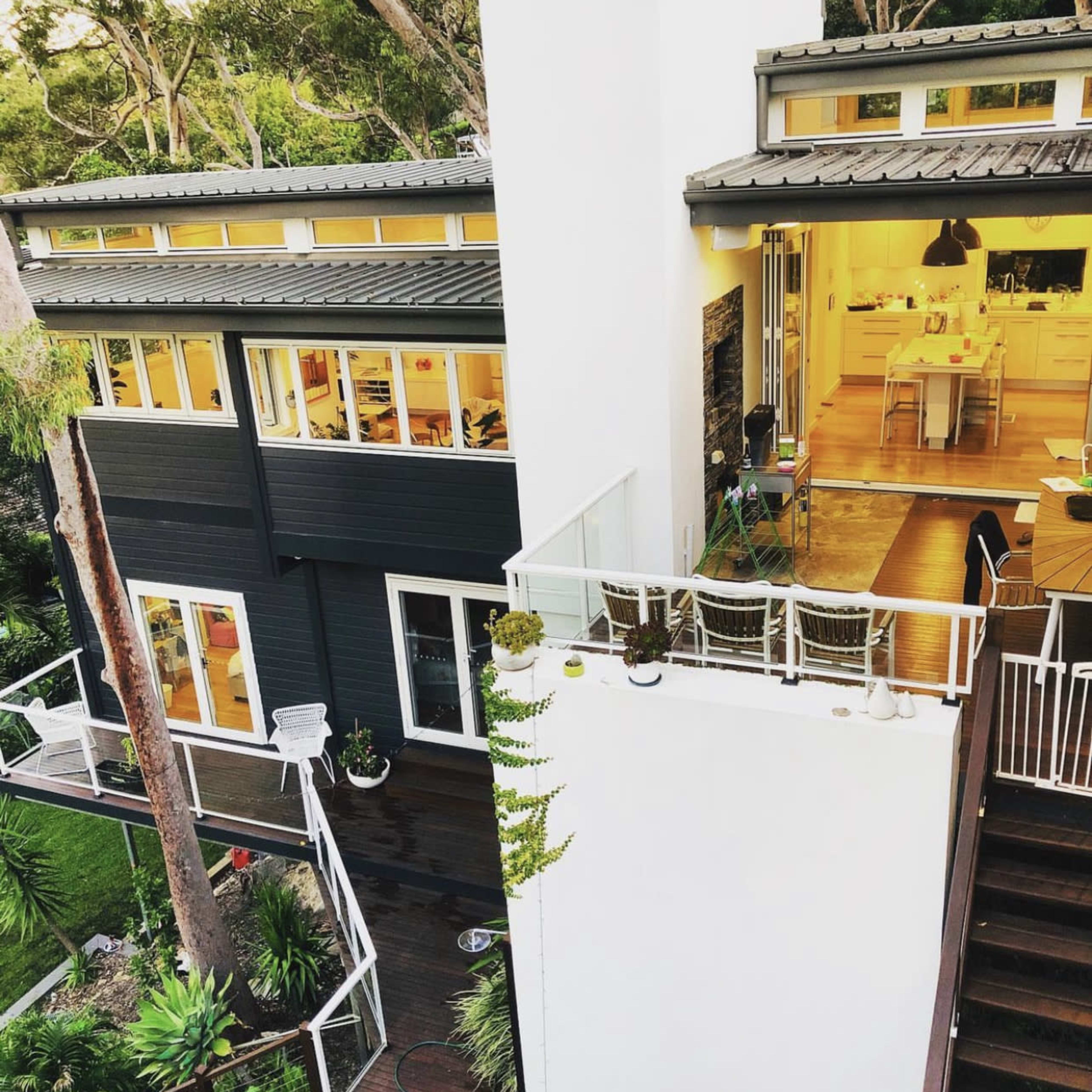 A modern two-story house with large windows surrounded by lush greenery, featuring a balcony and an outdoor seating area.