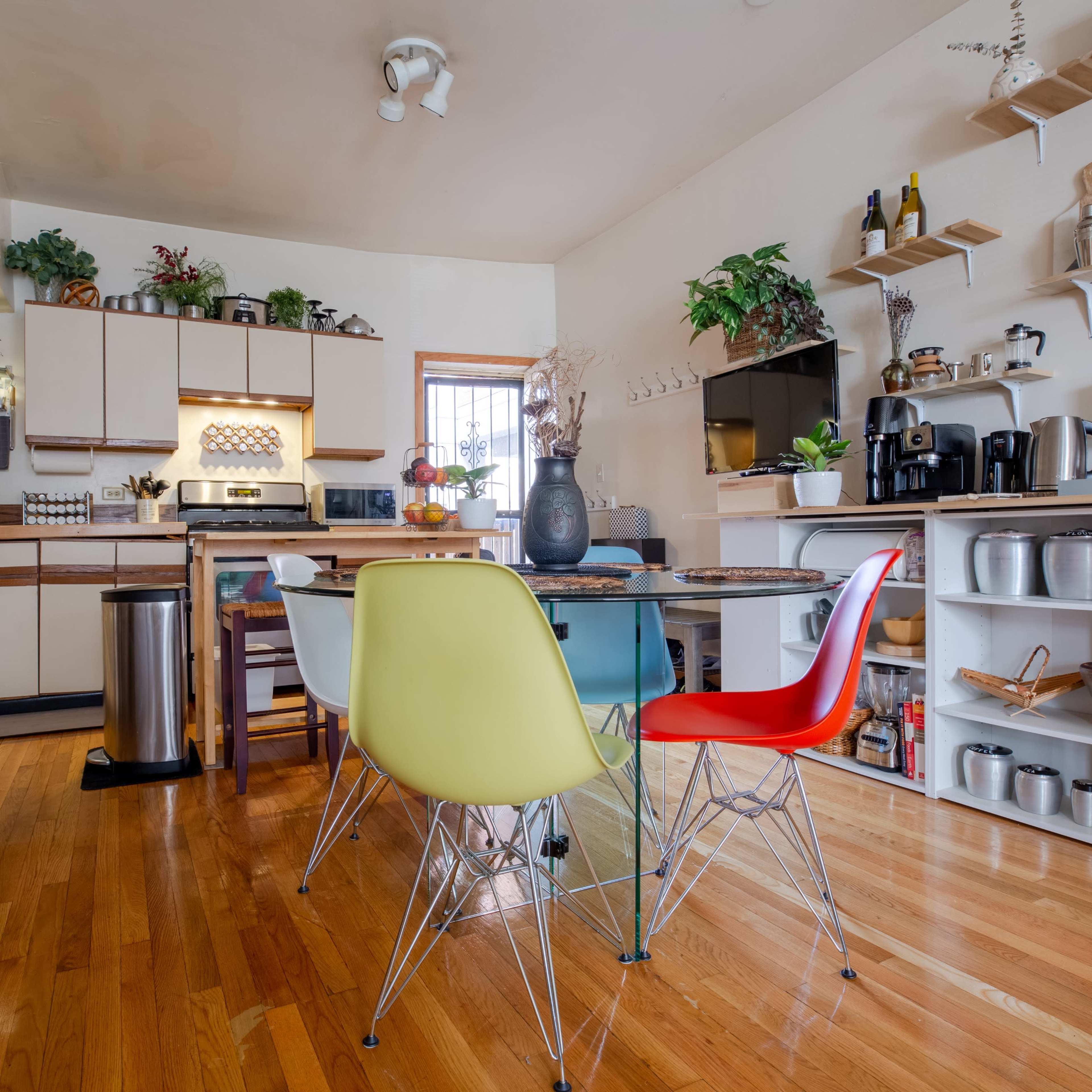 A well-organized kitchen with wooden floors, a dining table surrounded by colorful chairs, and shelves displaying various kitchen items.