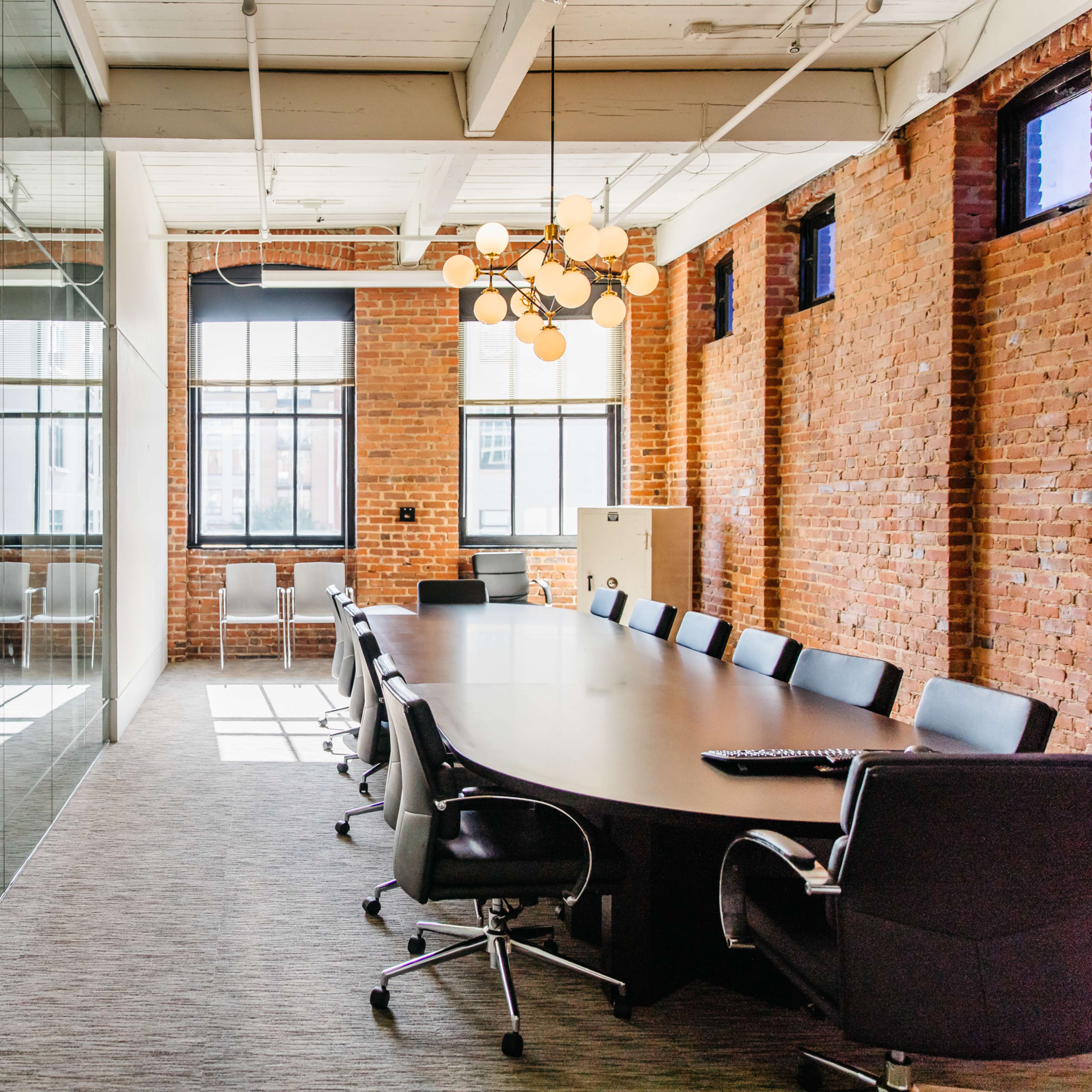 A conference room features a long table surrounded by black chairs, with a glass wall reflecting the space and brick walls in the background.