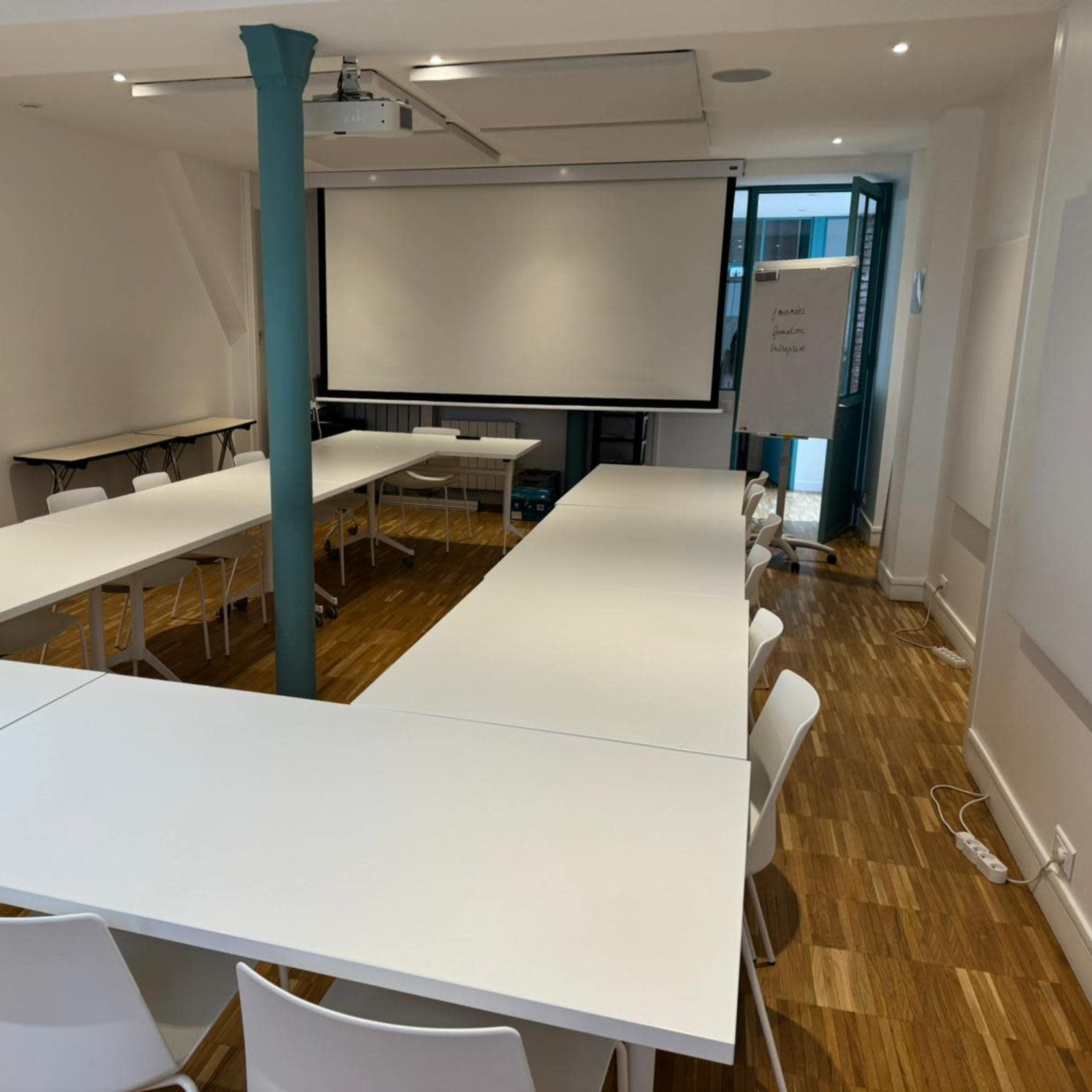 A meeting room with white tables arranged in a U-shape and several white chairs, featuring a projector screen and large windows.