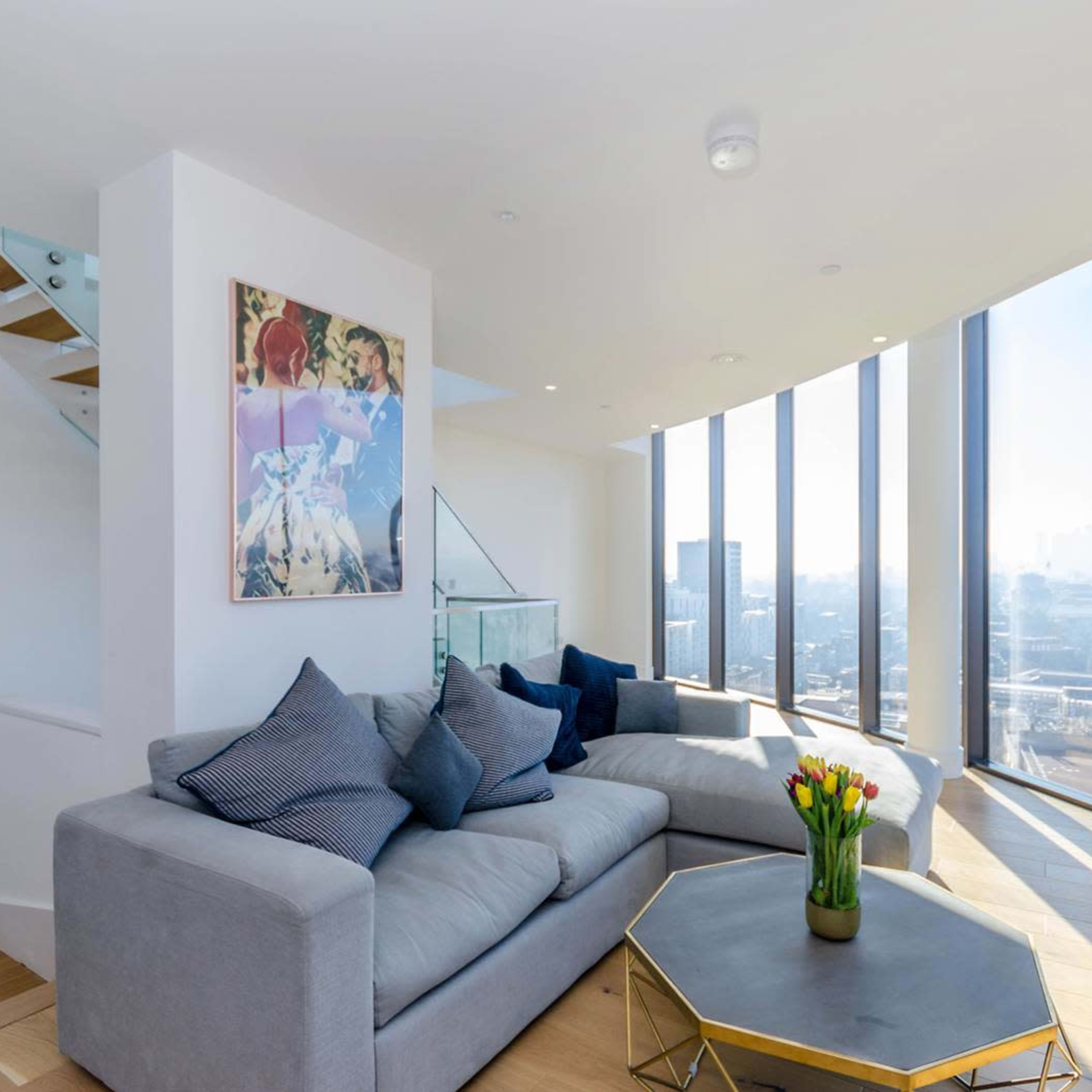A modern living room features a gray sectional sofa, a geometric coffee table, and large floor-to-ceiling windows offering a city view, with a colorful artwork on the wall.