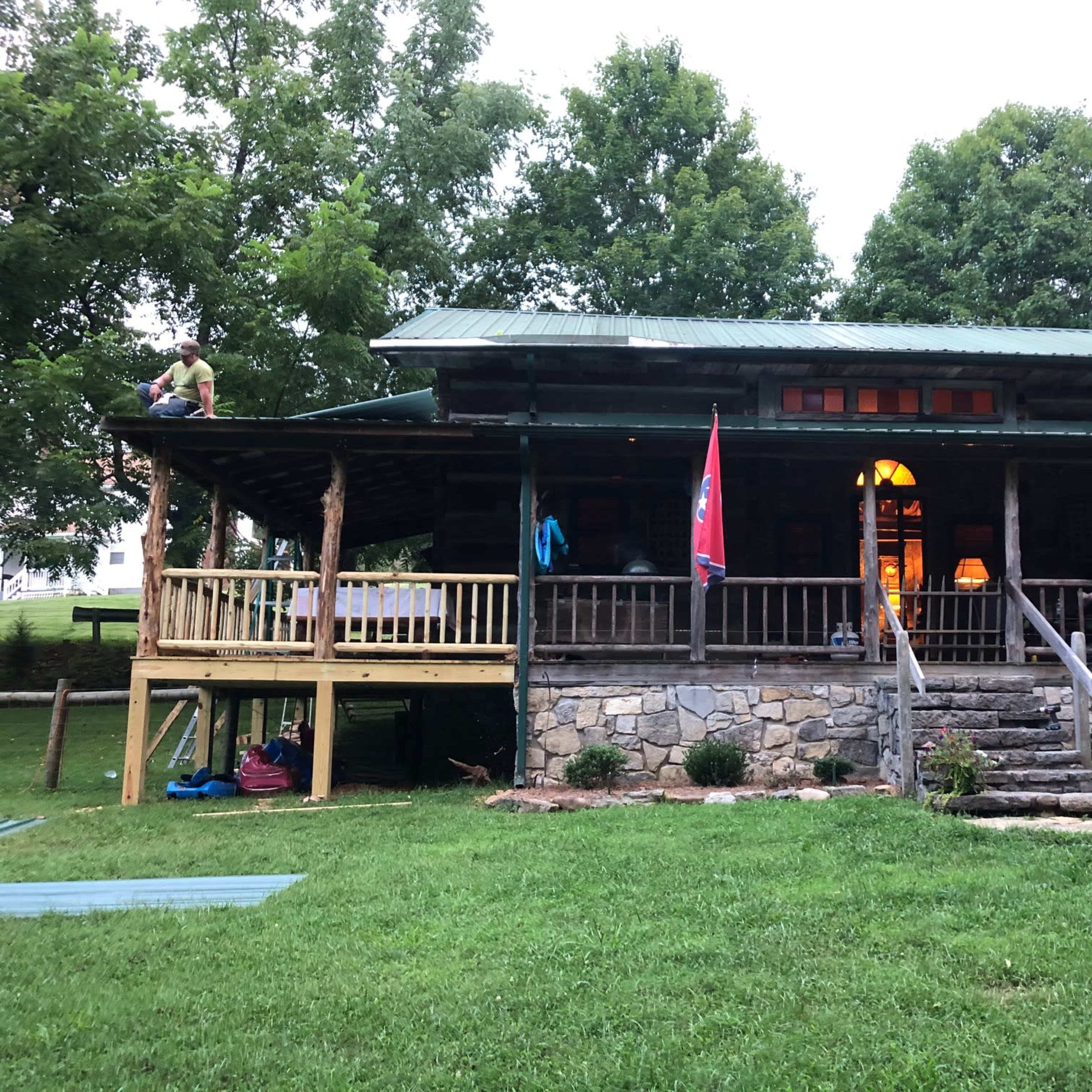 A wooden cabin with a green roof features a porch, stone steps, and flags from Texas and Tennessee, surrounded by grassy areas and trees.