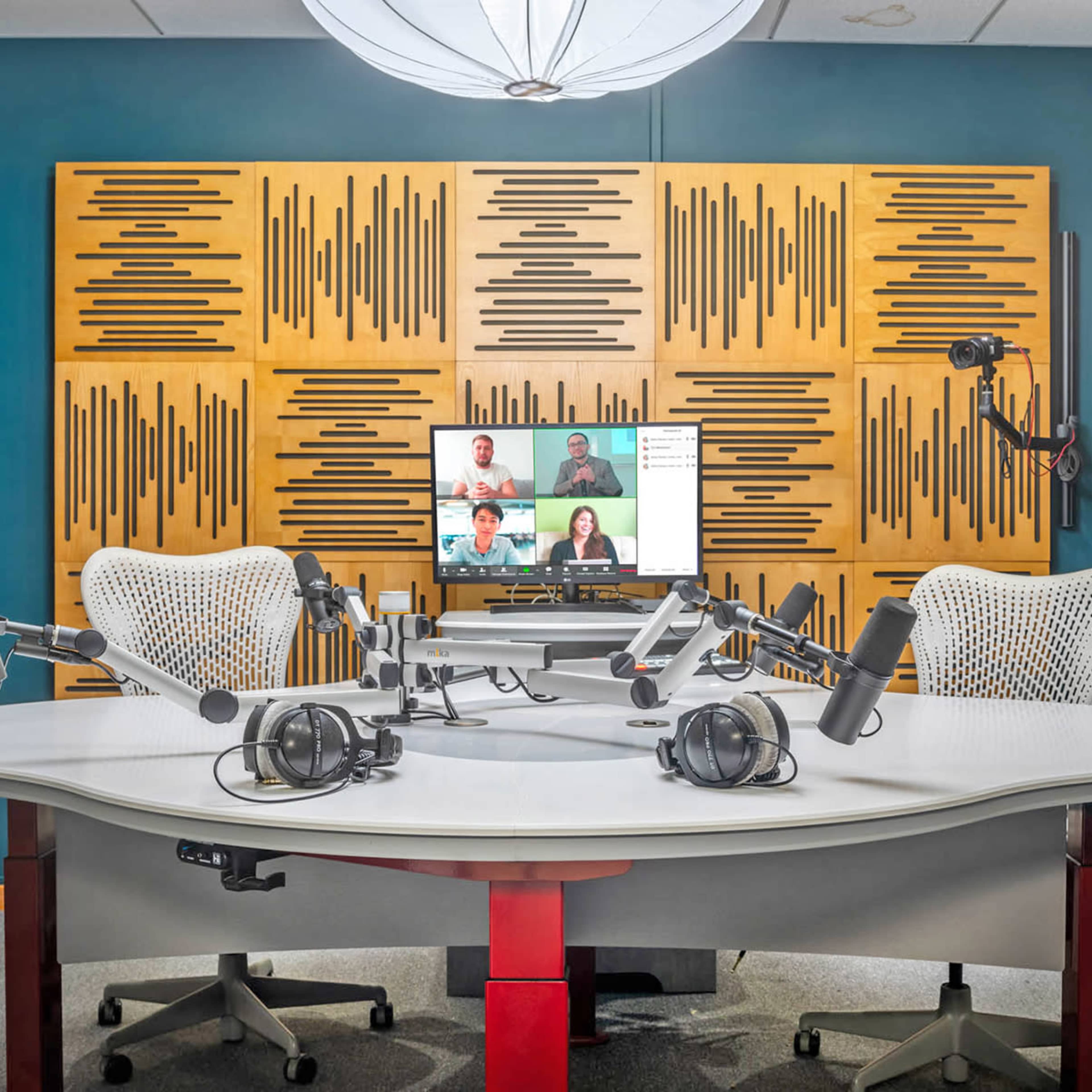 A modern recording studio with two chairs, multiple microphones, headphones, and a display screen showing remote participants.