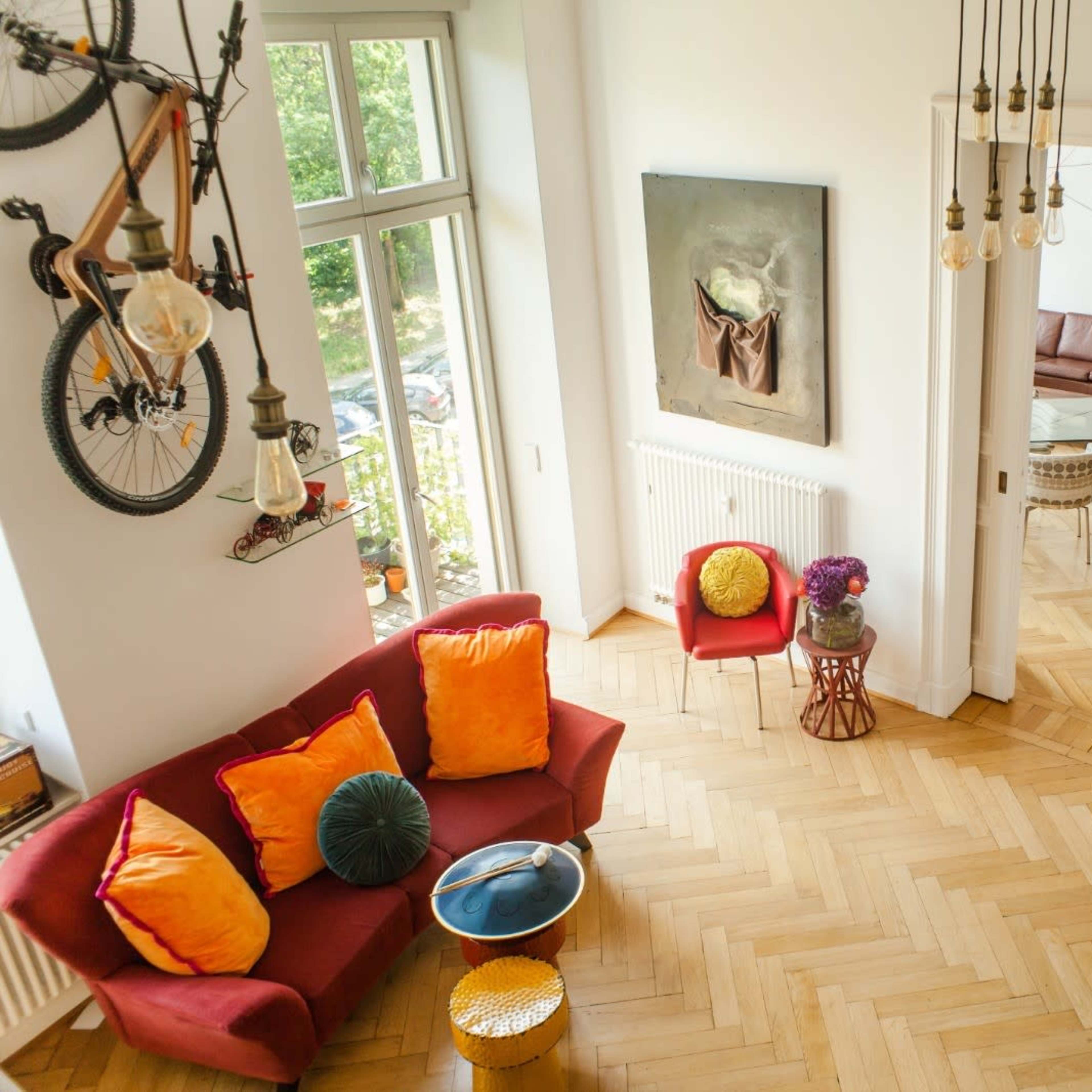 A bright living area features a red sofa with colorful cushions, a wooden floor, and a guitar on the wall, complemented by large windows and a dining space in the background.
