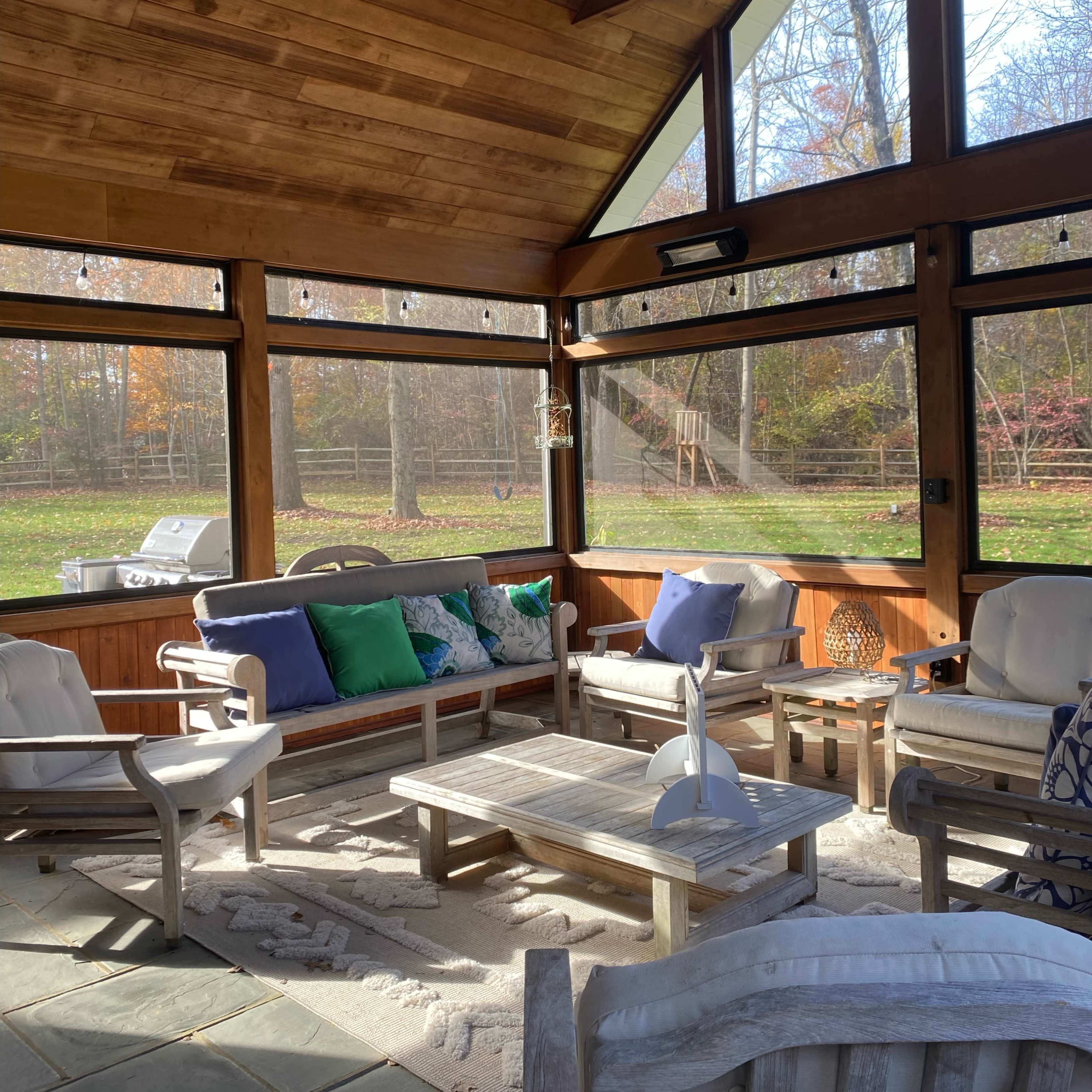 A cozy sunroom with wooden walls and ceiling, featuring comfortable seating arranged around a central coffee table, and large windows overlooking a green yard.