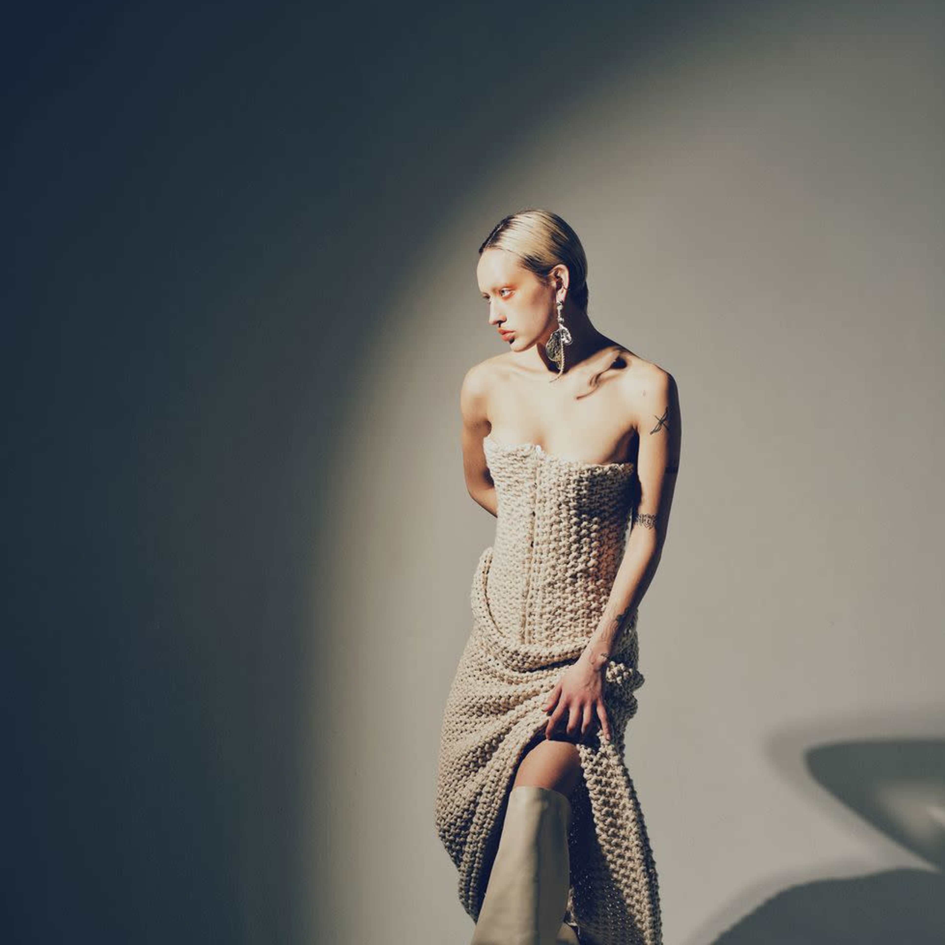 A model poses elegantly in a knitted, strapless dress and high-heeled boots against a softly lit background.