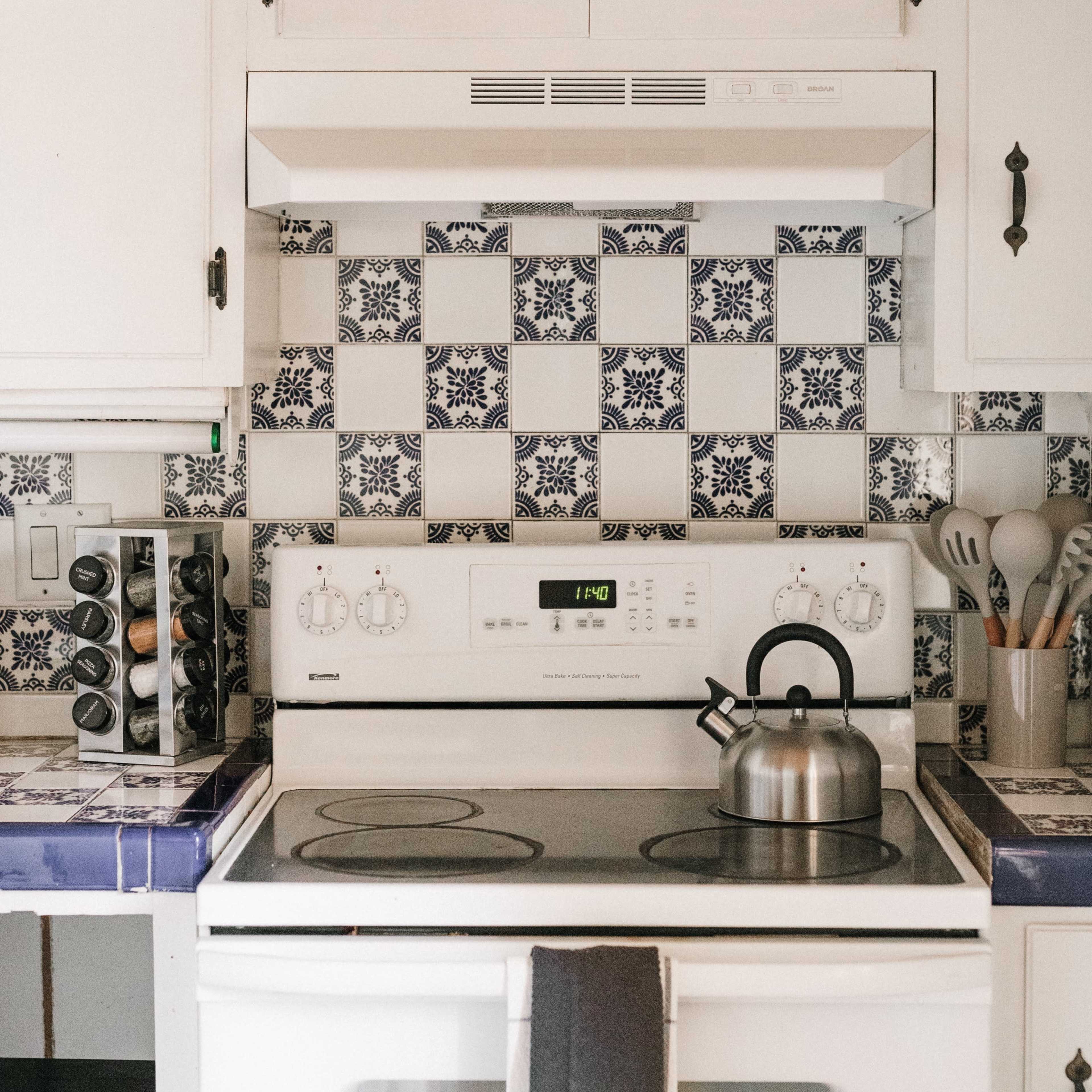 A kitchen with a white stove under a range hood, surrounded by blue and white patterned tiles, with a kettle on the stove and various cooking utensils displayed nearby.